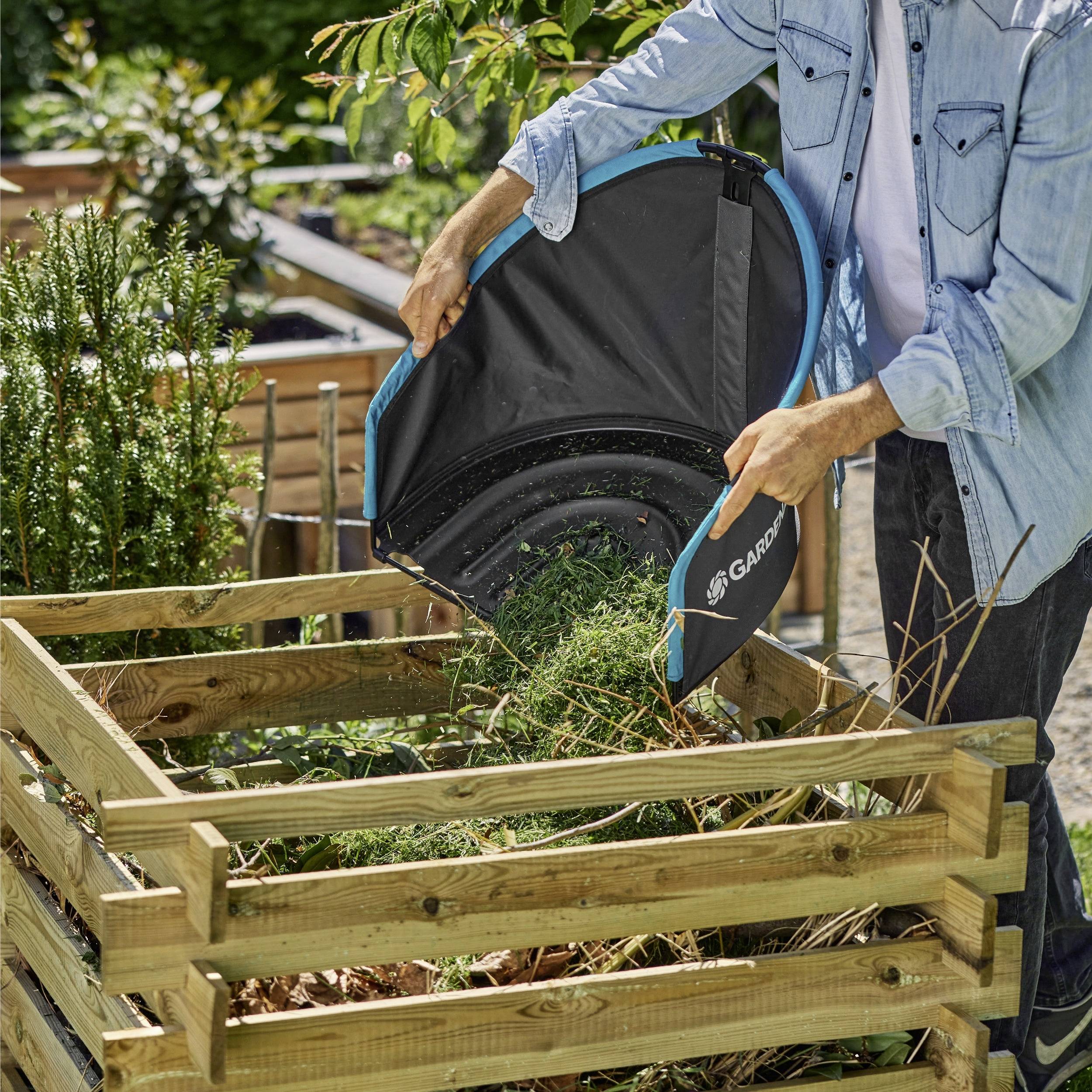 Une personne ajoute des déchets de jardin à un bac de compostage en utilisant un sac de jardin pliable. La scène se déroule dans un espace de jardin extérieur avec différentes plantes visibles.