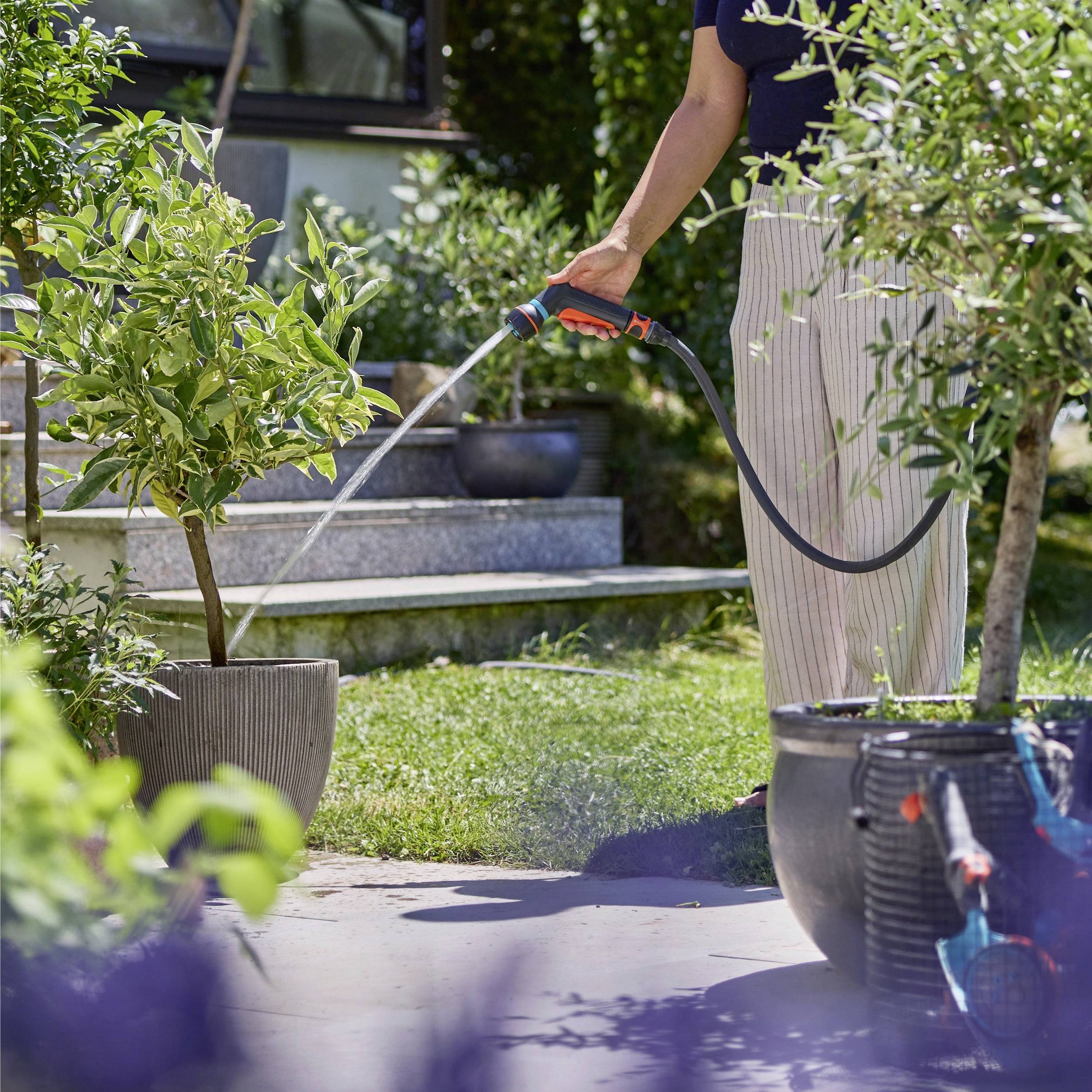 Un gros plan d'une main tenant un embout de tuyau d'arrosage, projetant de l'eau sur une terrasse, avec de la végétation floue en arrière-plan.