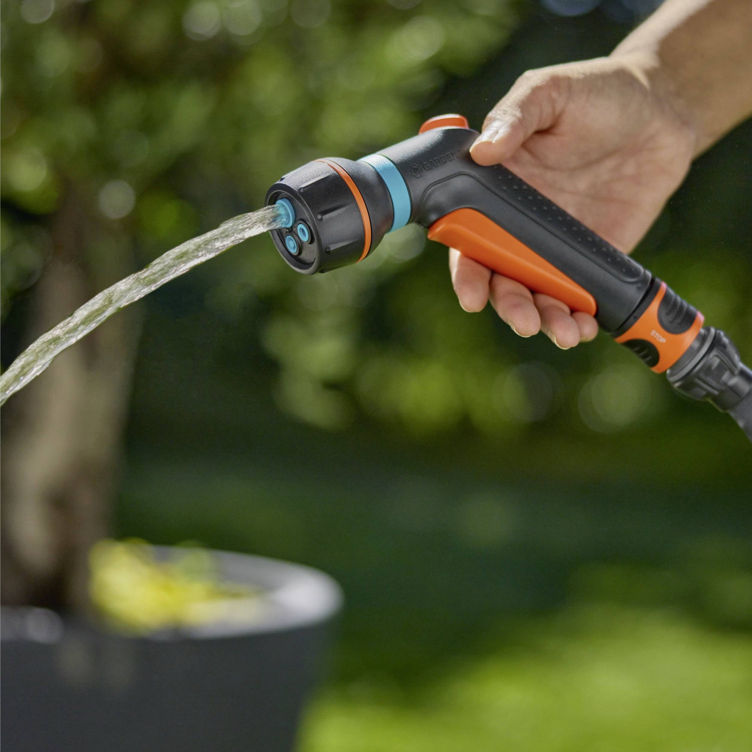 Une main tient un embout de tuyau d'arrosage orange et bleu, projetant de l'eau avec un pot de plante visible dans un arrière-plan flou.