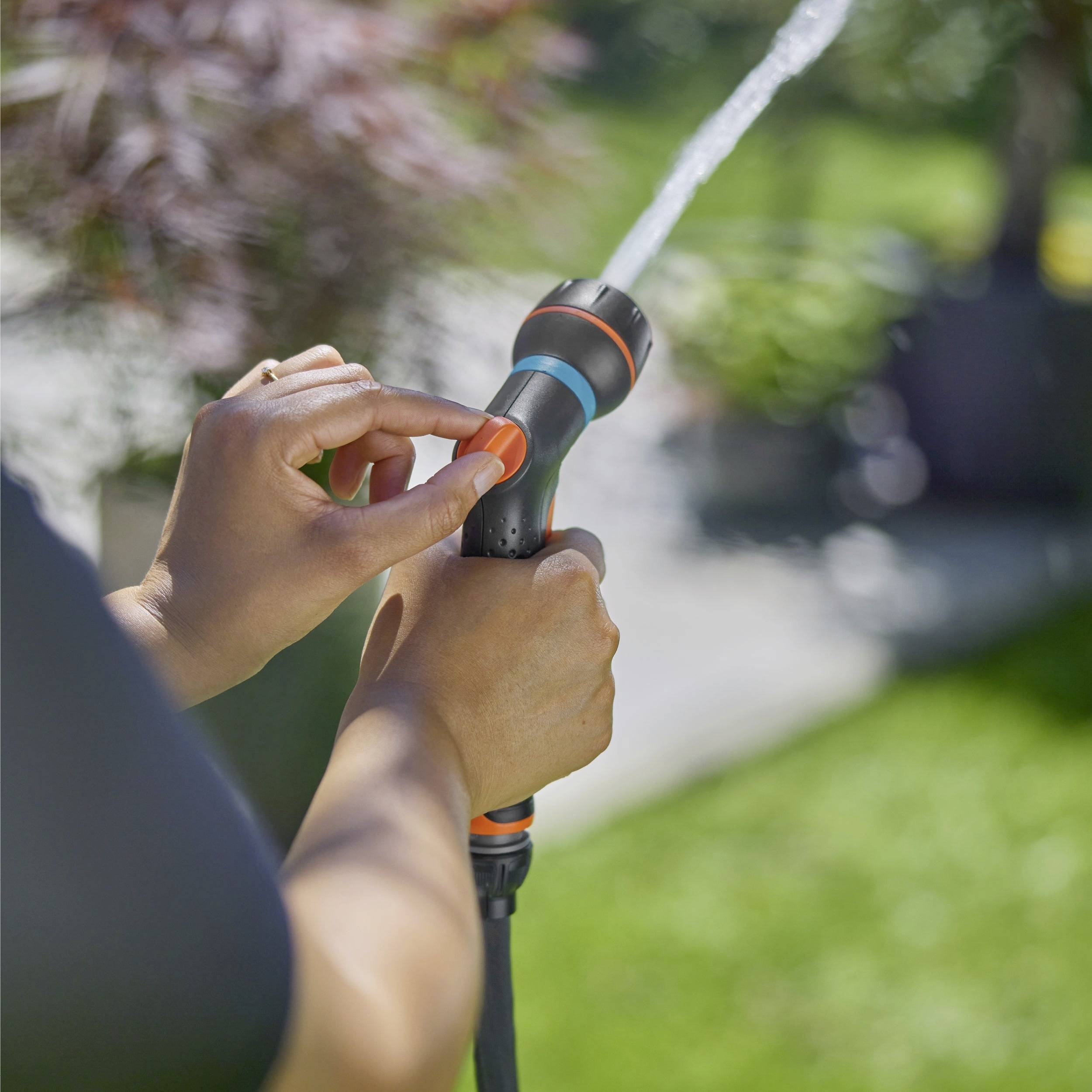 Un individu ajustant un pistolet d'arrosage sur un tuyau d'arrosage, projetant de l'eau dans un jardin ensoleillé avec un arrière-plan d'herbe.
