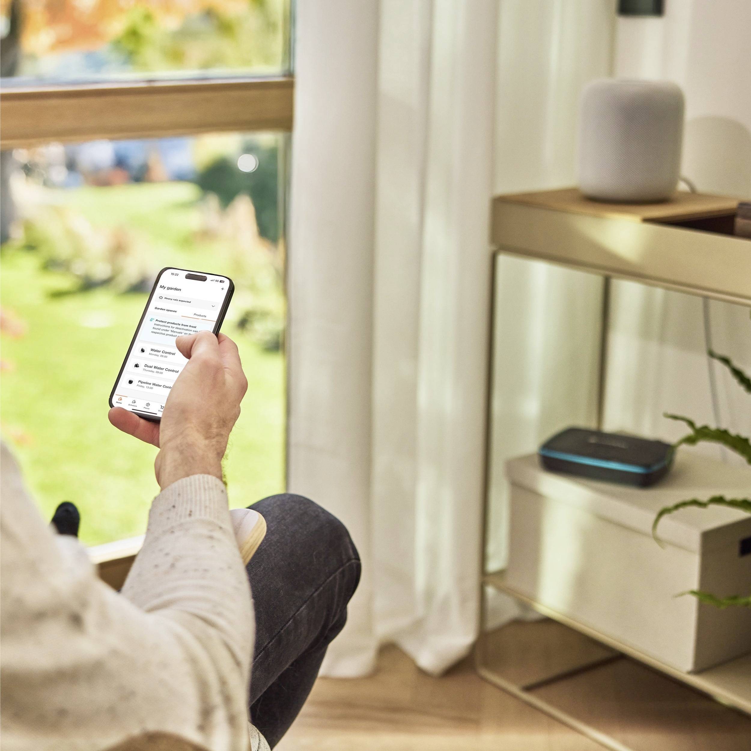 Une personne est assise à l'intérieur, tenant un smartphone et consultant des notifications. À proximité, un haut-parleur intelligent et un routeur sont posés sur une étagère.