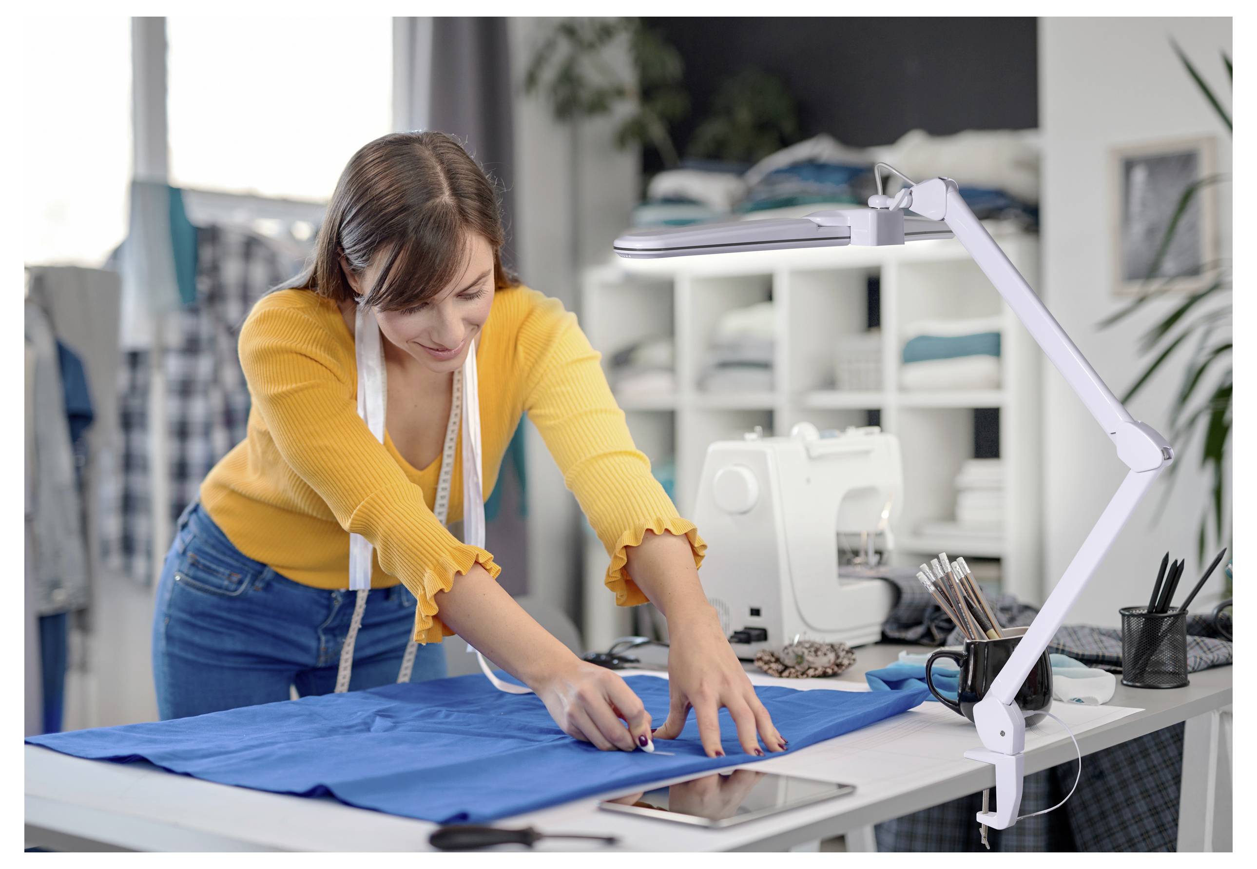 Une personne mesure du tissu avec un mètre ruban dans une salle de couture, entourée d'outils de couture et d'une machine à coudre. Elle semble concentrée sur son travail.