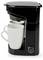 Une cafetière noire avec une tasse blanche positionnée sous le distributeur, prête à être utilisée. Le bouton marche/arrêt est visible sur le socle.