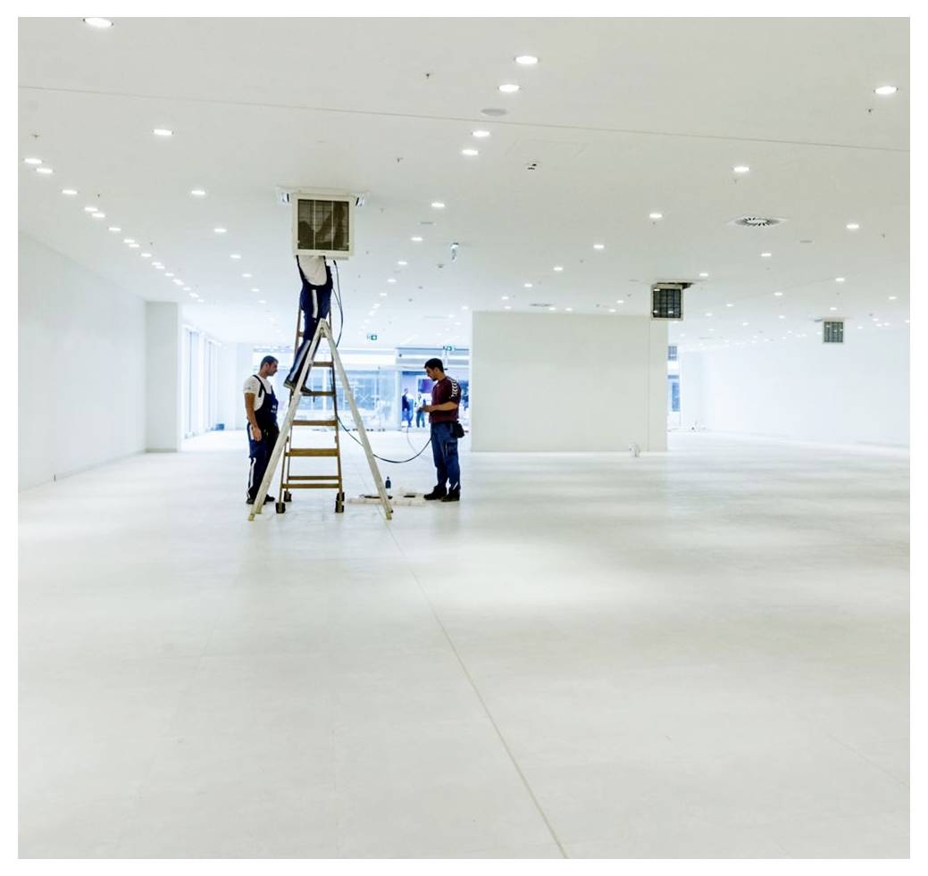 Deux ouvriers se tiennent sur et autour d'une échelle dans une grande pièce blanche, vide et bien éclairée, semblant effectuer une maintenance sur un luminaire au plafond.