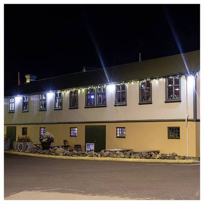 Un long bâtiment de nuit, éclairé par des lumières extérieures, présente des pierres décoratives le long de sa base et de petites fenêtres aux encadrements rouges.