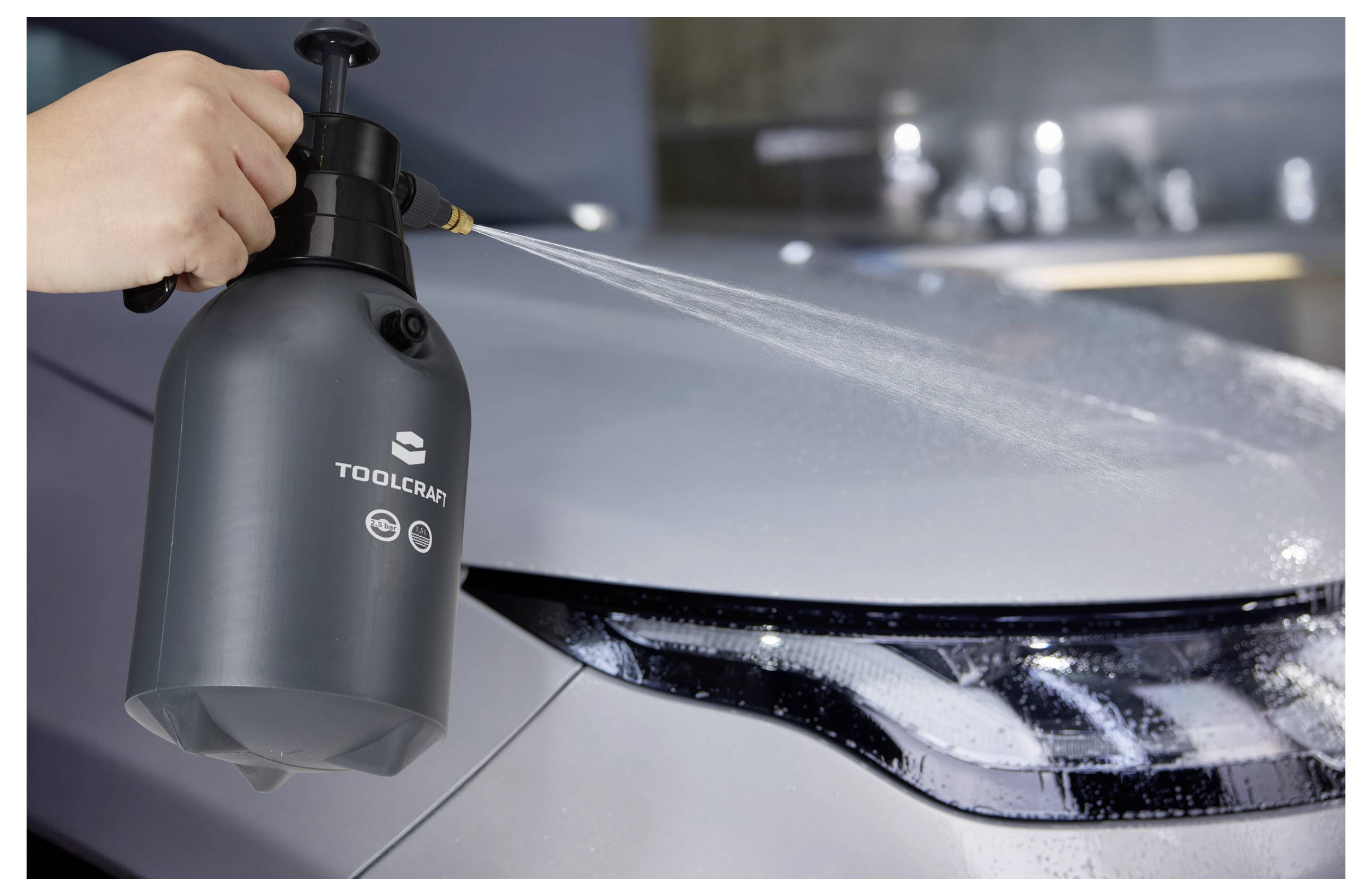 Une personne pulvérise de l'eau sur un phare de voiture à l'aide d'une bouteille noire étiquetée « Toolcraft », mettant en évidence le processus de nettoyage de la voiture.