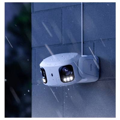 Une caméra de sécurité fixée sur un mur, capturant une nuit pluvieuse. La caméra possède deux objectifs et des lumières LED, avec des gouttelettes d'eau visibles.