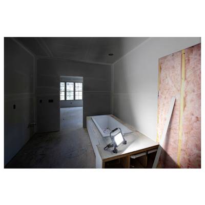 Une pièce faiblement éclairée en cours de rénovation, présentant une baignoire inachevée entourée de matériaux de construction, avec un projecteur de chantier illuminant la scène.