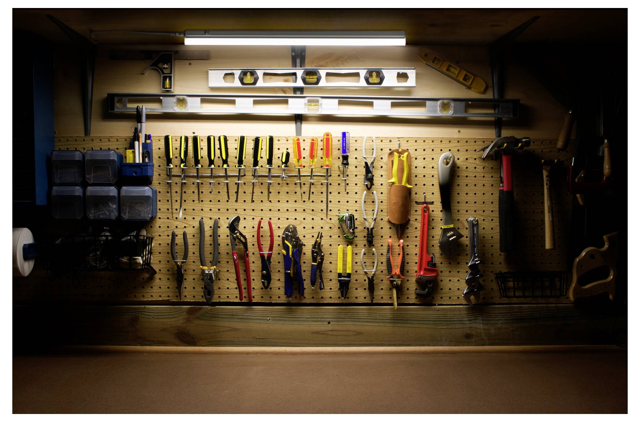 Un panneau perforé dans un atelier faiblement éclairé présente des outils à main soigneusement organisés, comprenant des pinces, des tournevis et des marteaux, avec des niveaux posés au-dessus.
