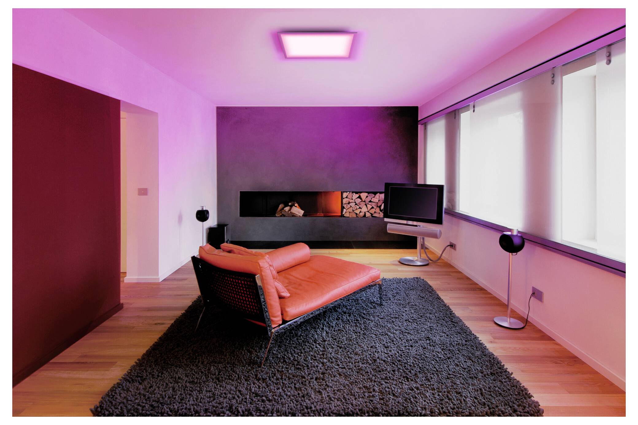 Un salon moderne avec une chaise rouge sur un tapis gris, un éclairage violet, une télévision murale et une cheminée intégrée avec un espace de rangement pour le bois.