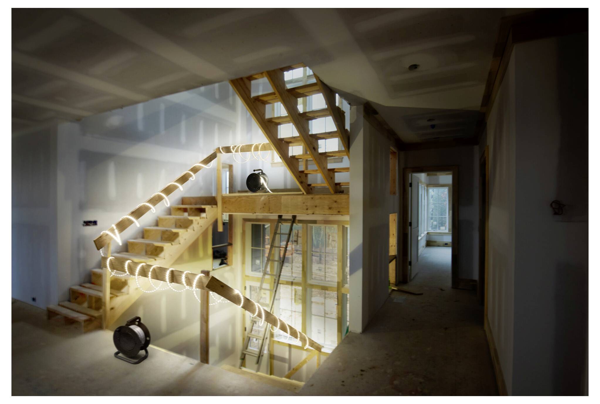 Un escalier en bois en cours de construction avec une échelle à côté, éclairé par des projecteurs de chantier dans une pièce inachevée montrant des plaques de plâtre et des ouvertures de portes.