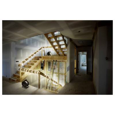 Un escalier en bois en cours de construction avec une échelle à côté, éclairé par des projecteurs de chantier dans une pièce inachevée montrant des plaques de plâtre et des ouvertures de portes.