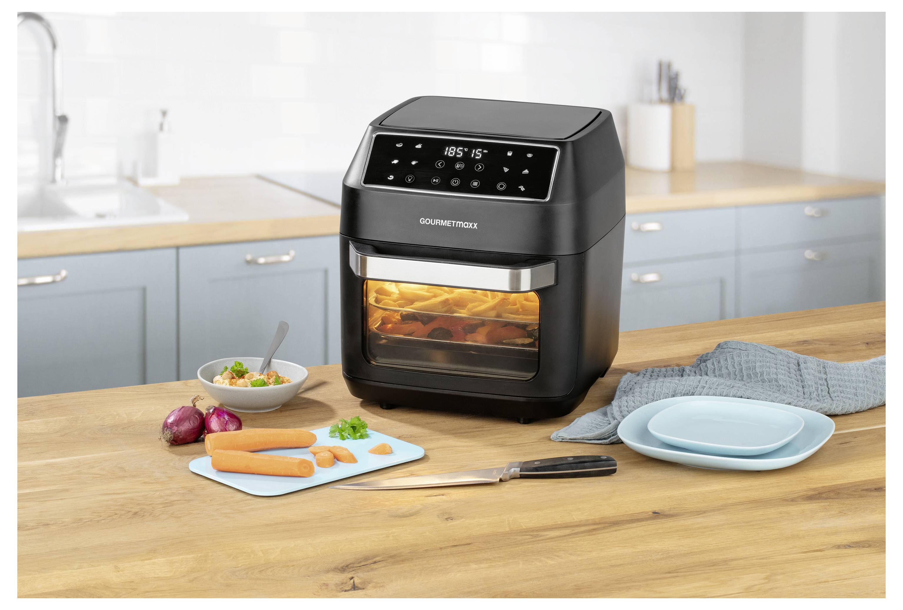 Un air fryer numérique noir sur un comptoir de cuisine est en train de cuire du poulet. À proximité se trouvent des légumes coupés, une planche à découper avec des carottes, un couteau et des assiettes bleues.
