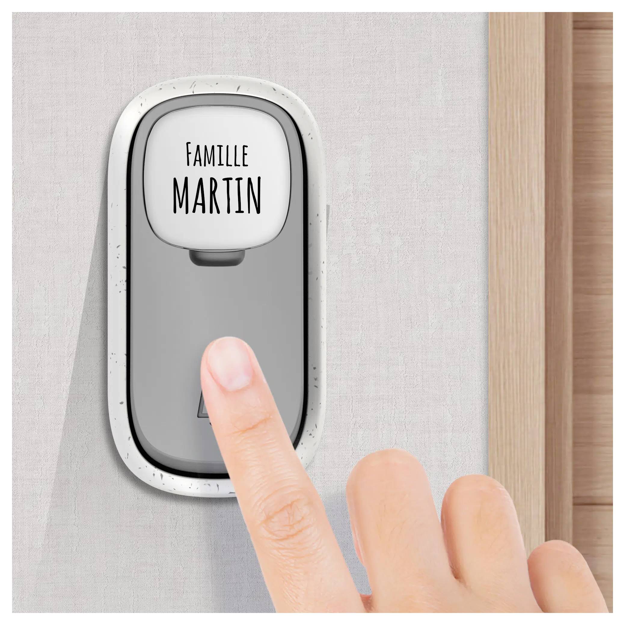 Un doigt d'une personne appuie sur une sonnette étiquetée « Famille Martin » sur un mur, à côté d'un cadre en bois.