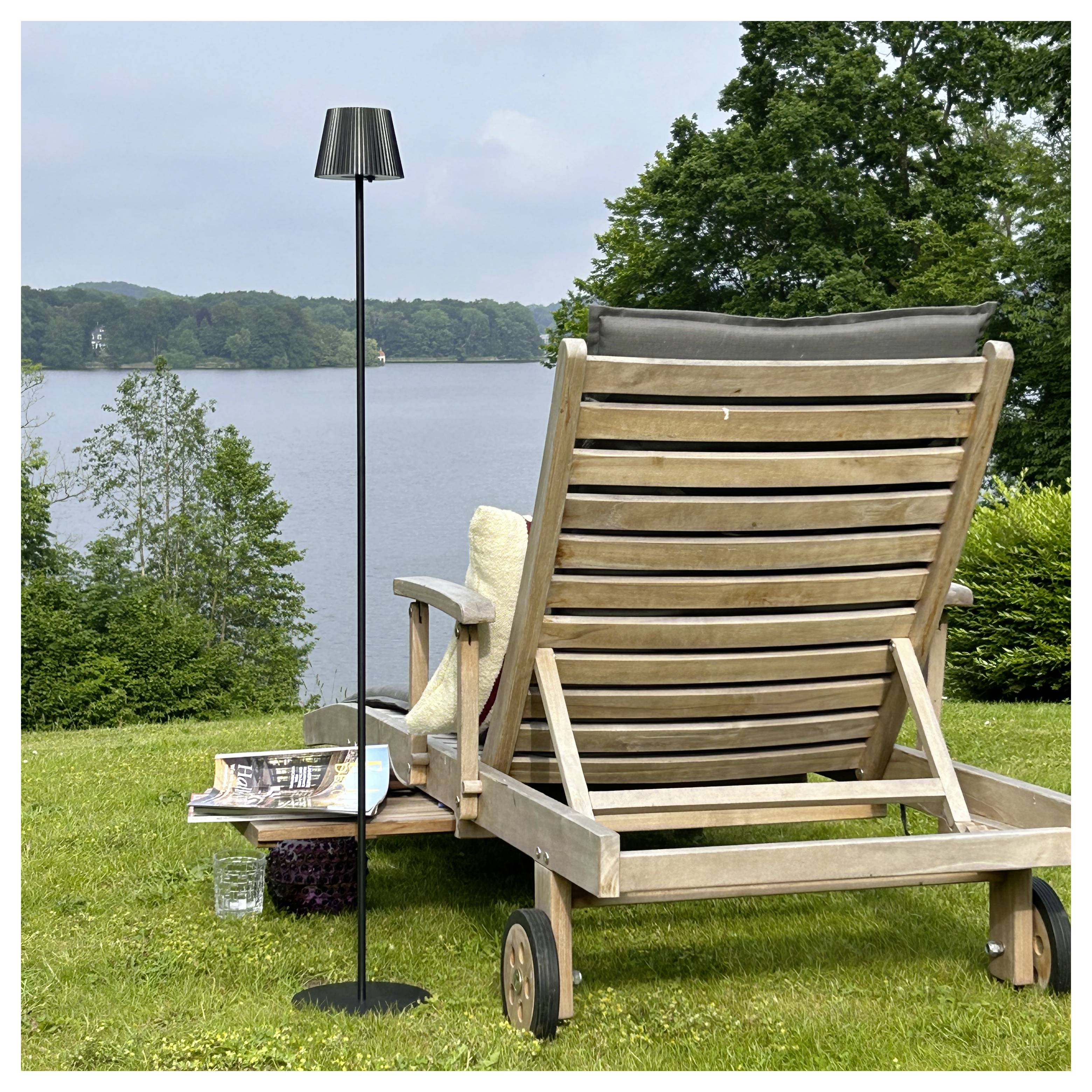 Une chaise longue en bois avec un coussin et une petite table supportant des magazines et des raisins surplombe un lac pittoresque et des arbres.