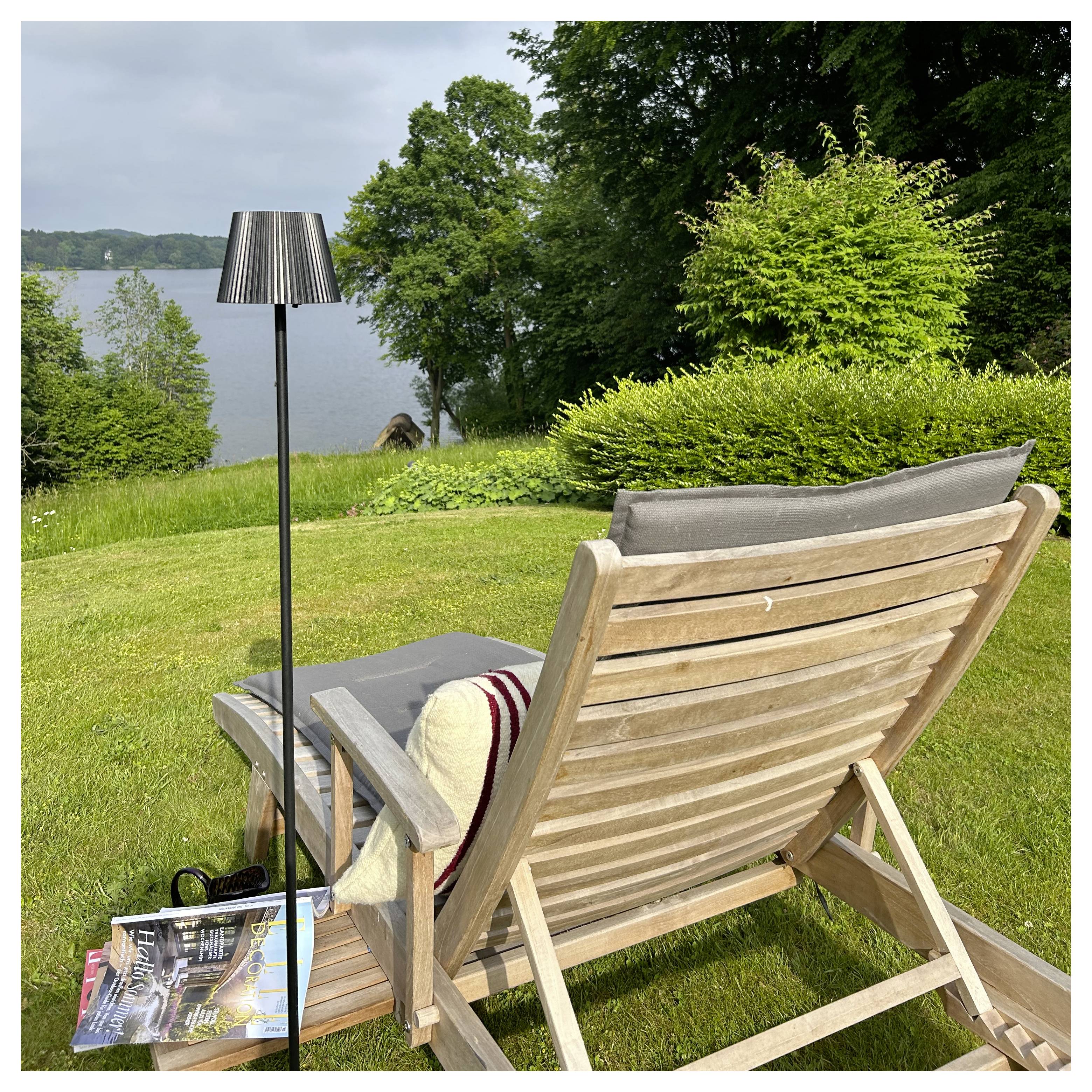 Une chaise longue en bois avec un coussin et une couverture fait face à une vue sur le lac, entourée de verdure luxuriante et avec une lampe à ses côtés.