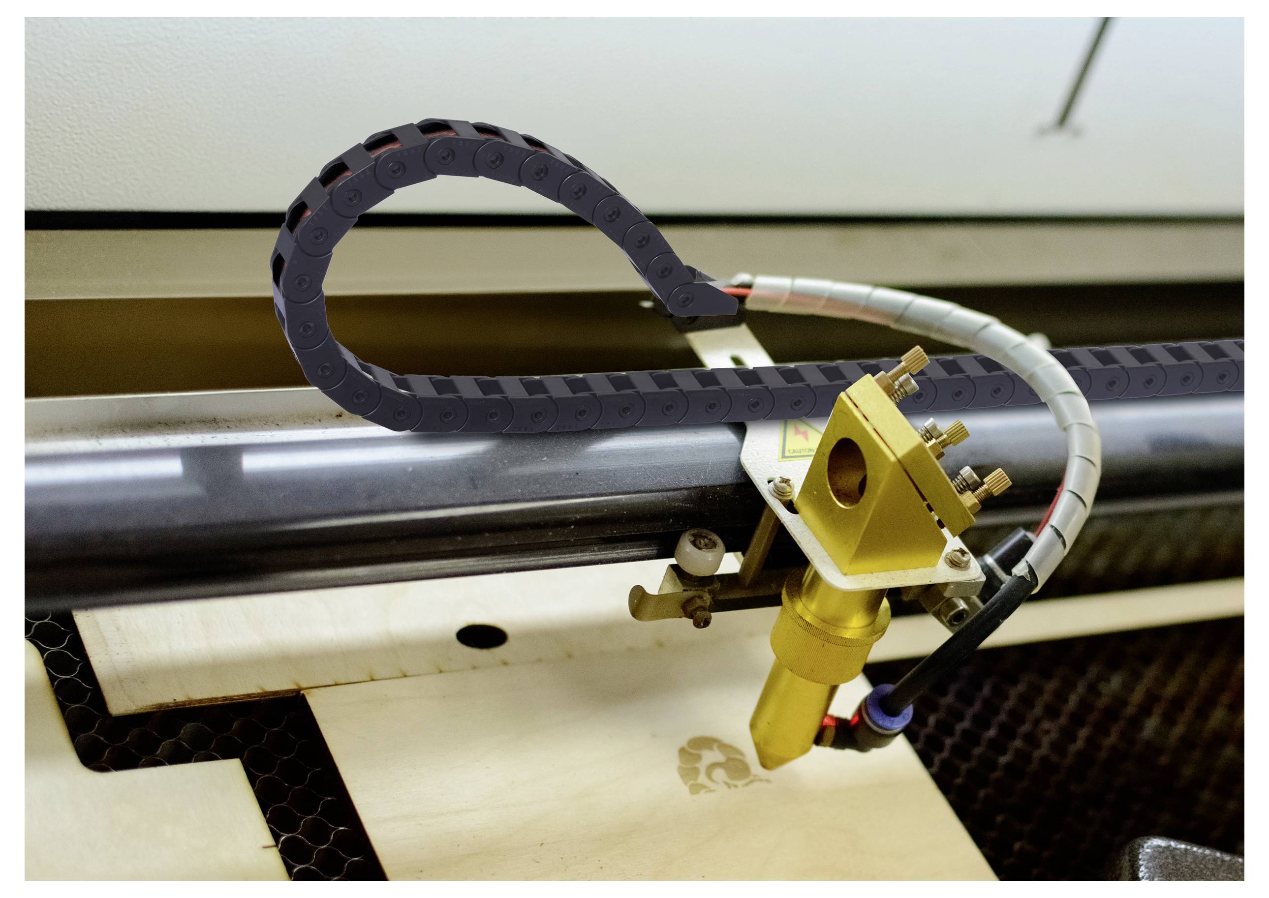 Machine de découpe laser gravant un logo sur une feuille de bois, avec la tête de coupe et le câble flexible visibles au-dessus de la surface de travail.