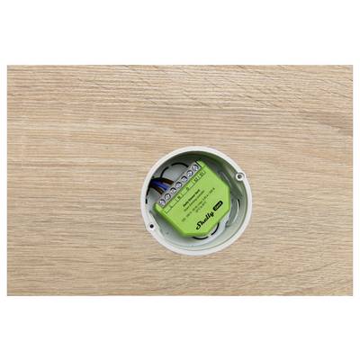 Un appareil électrique vert est installé dans une prise murale circulaire. Il est étiqueté « Shelly 1 ». Le mur environnant a une texture de bois clair.