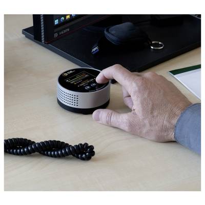 Une main appuie sur un petit appareil électronique rond posé sur un bureau, avec un moniteur d'ordinateur et des accessoires en arrière-plan.