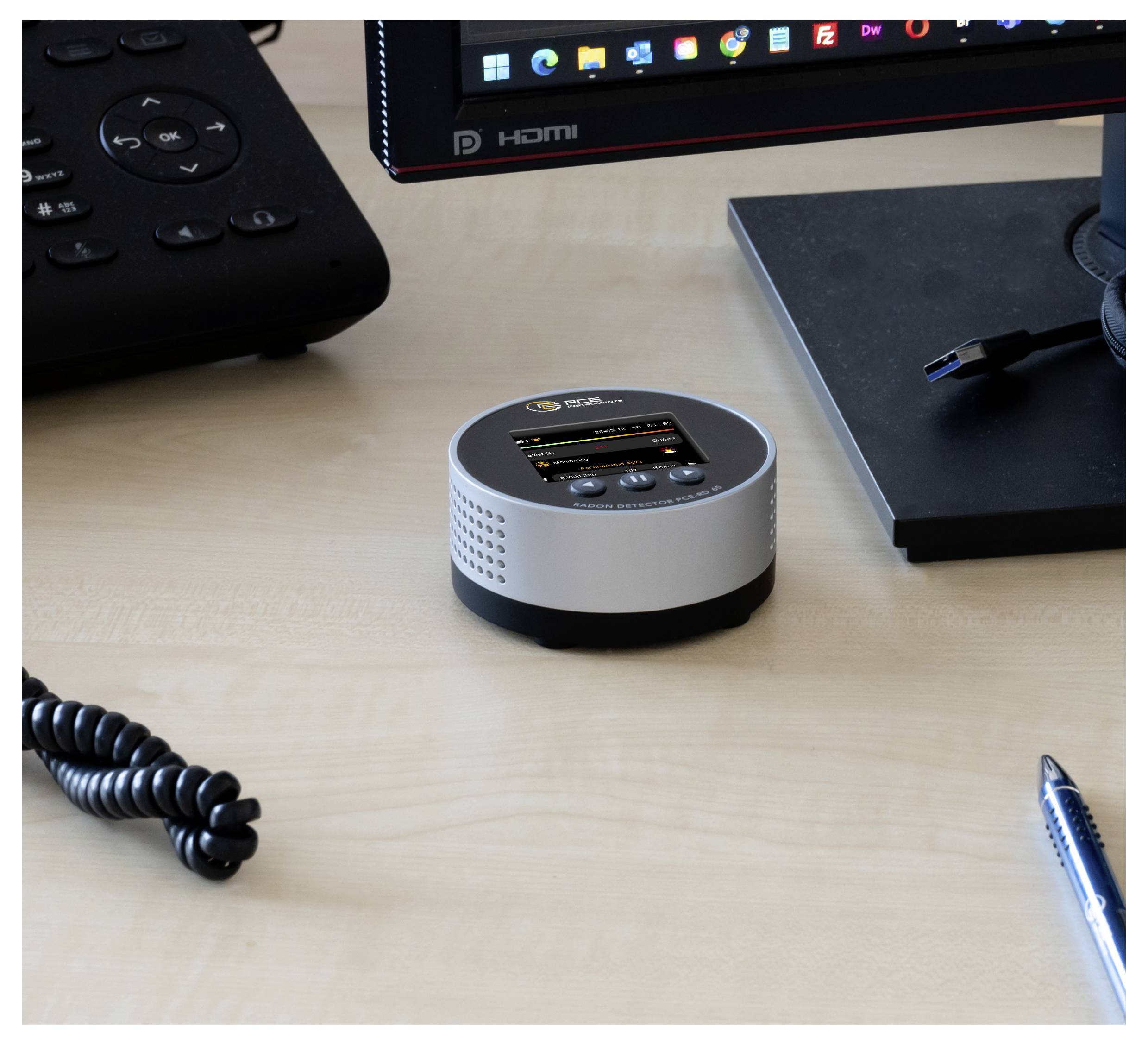 Petit appareil de table rond en argent avec un minuscule écran et des boutons sur un bureau à côté d'un moniteur, une télécommande, un stylo et un câble.