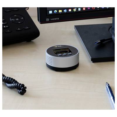 Petit appareil de table rond en argent avec un minuscule écran et des boutons sur un bureau à côté d'un moniteur, une télécommande, un stylo et un câble.