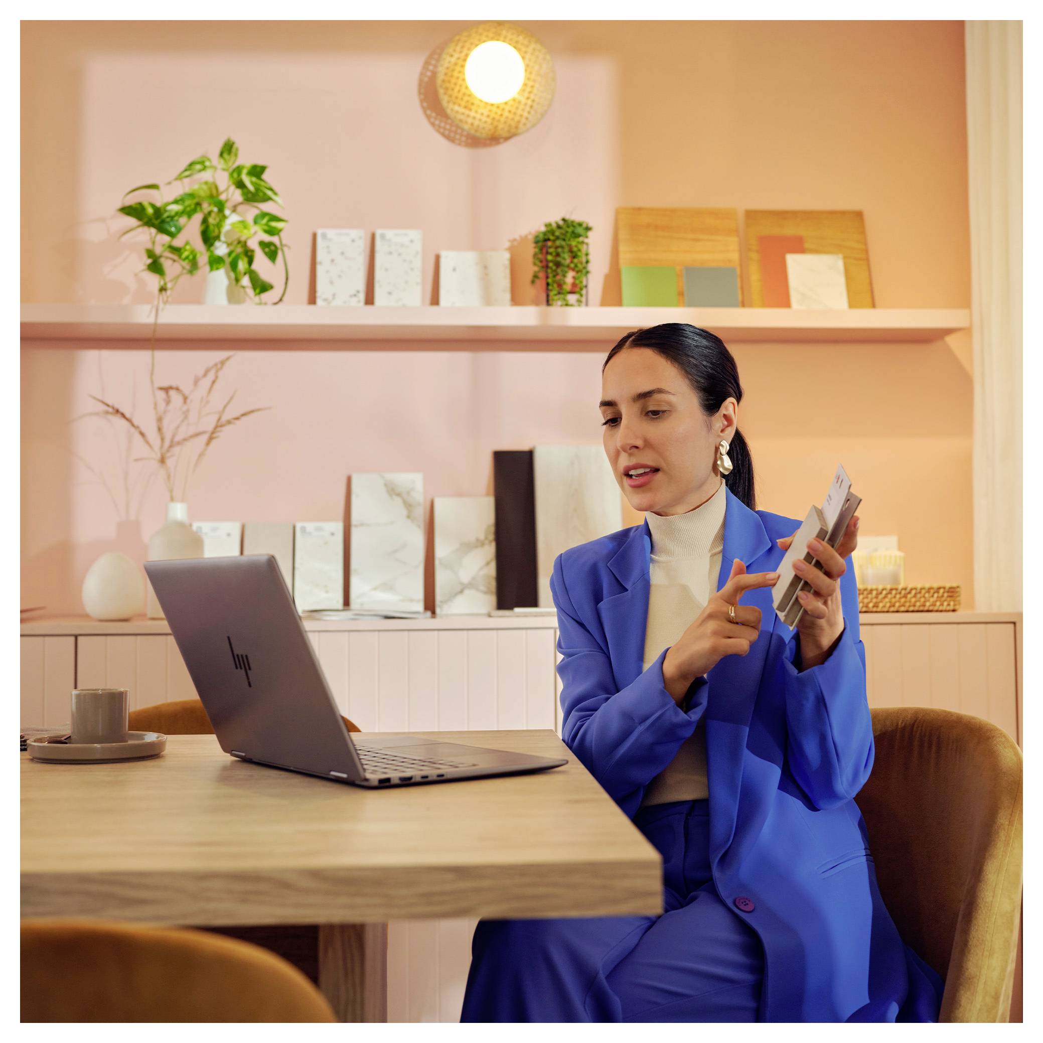 Une personne en costume bleu interagit avec un ordinateur portable à une table en bois, entourée d'objets décoratifs et de plantes sur des étagères roses.
