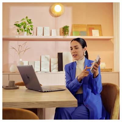 Une personne en costume bleu interagit avec un ordinateur portable à une table en bois, entourée d'objets décoratifs et de plantes sur des étagères roses.