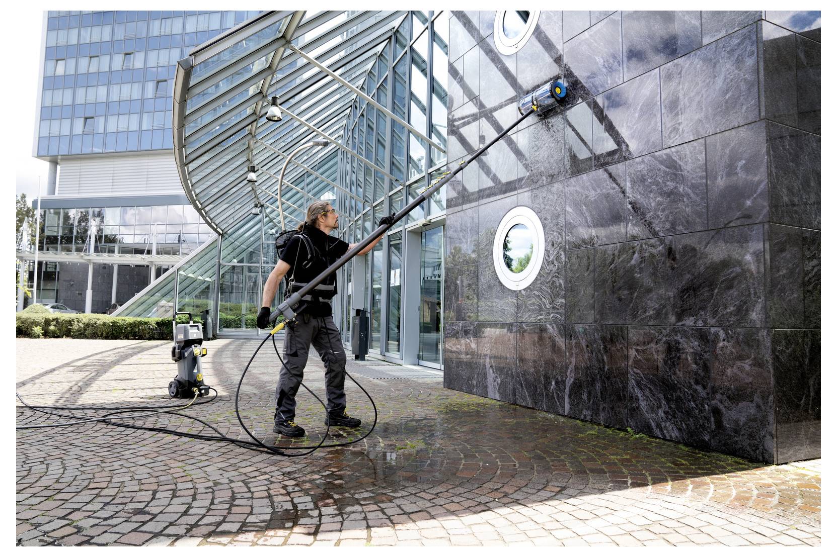 Un travailleur utilise un nettoyeur haute pression sur un bâtiment moderne et élevé en verre et en pierre, avec de l'eau et un tuyau de nettoyage visibles.