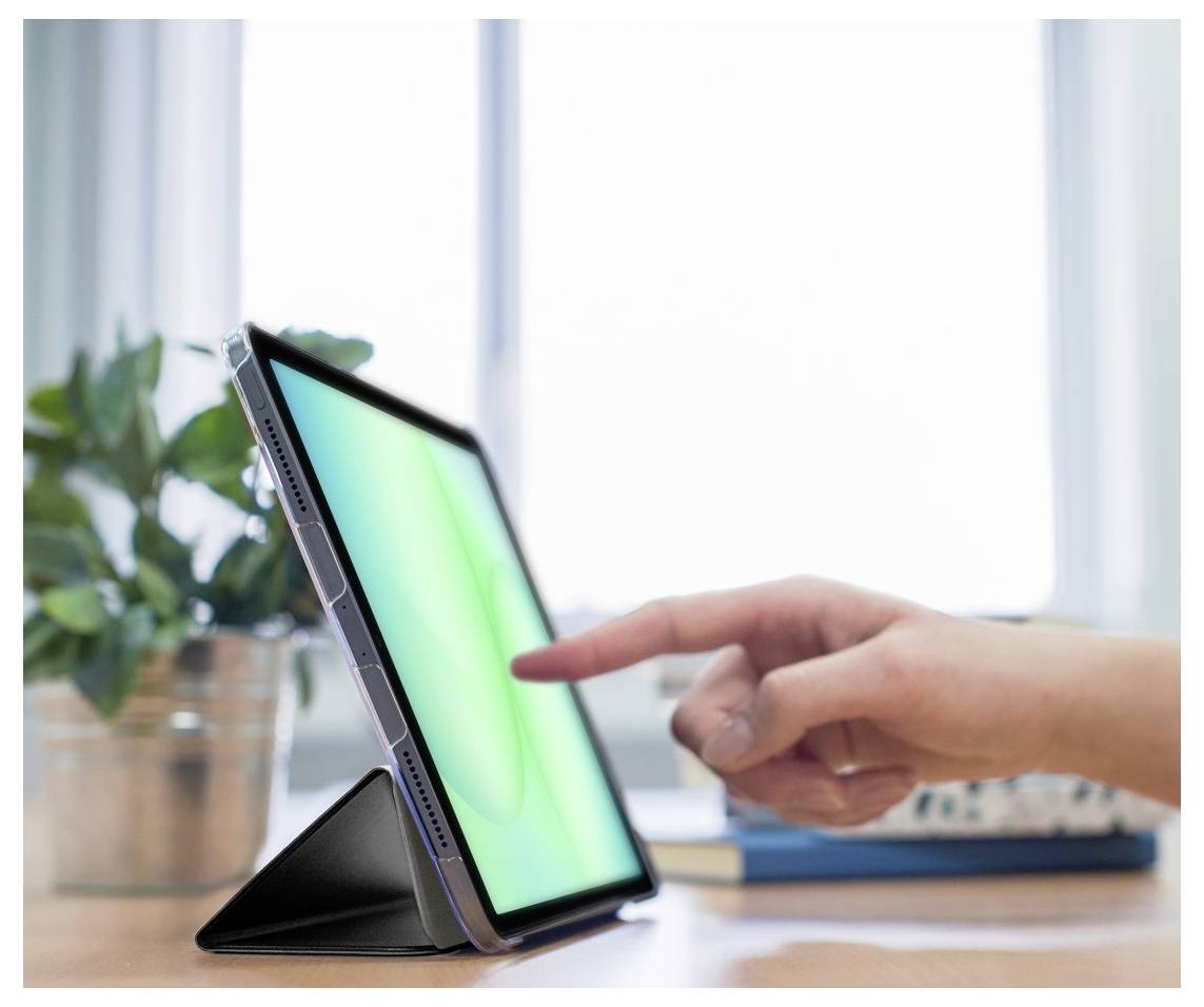 Une main touche l'écran d'une tablette, qui repose sur un support. Une plante et un carnet sont visibles dans l'arrière-plan flou.