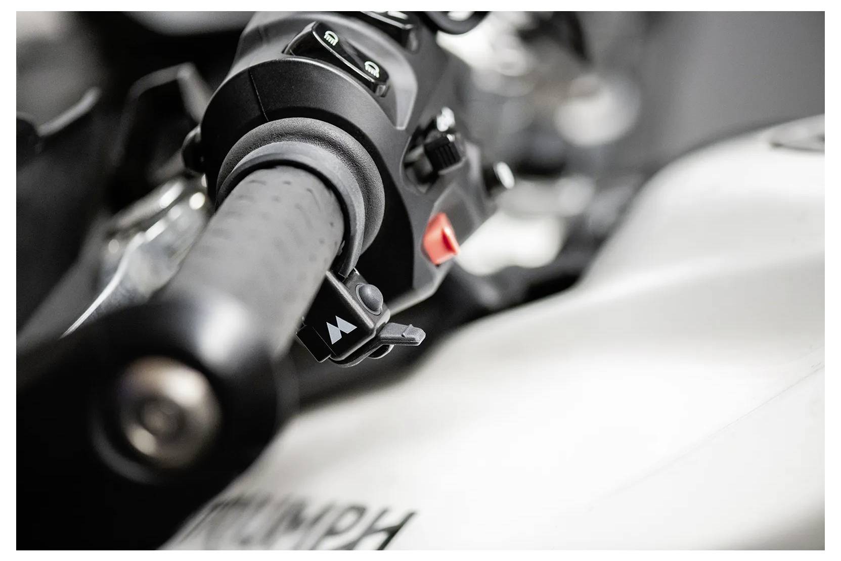 Gros plan d'un guidon de motocyclette montrant la poignée, certaines commandes, et une partie du réservoir de carburant avec des lettres floues.