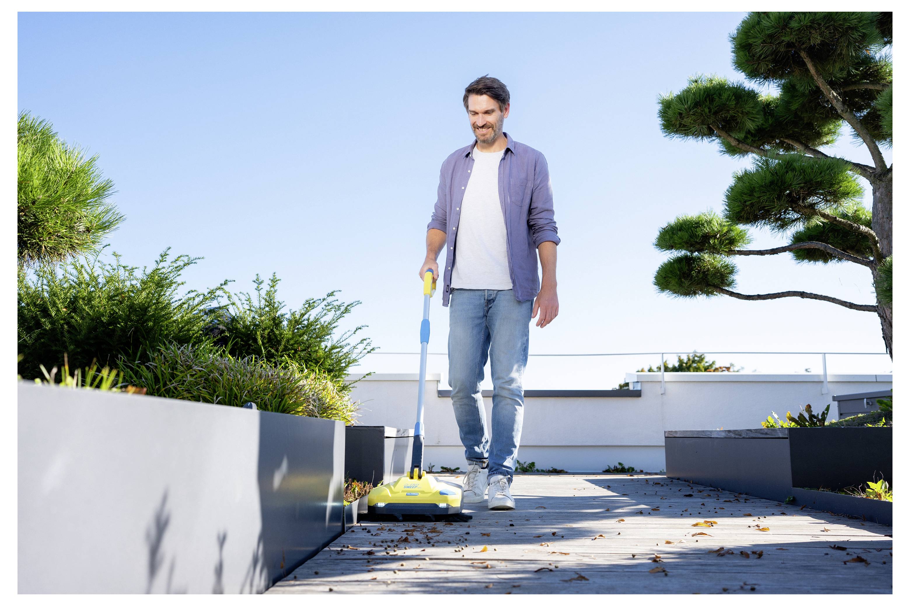 Une personne nettoie un jardin sur toit moderne avec un balai électrique à long manche. Une verdure luxuriante et un ciel bleu clair en arrière-plan.