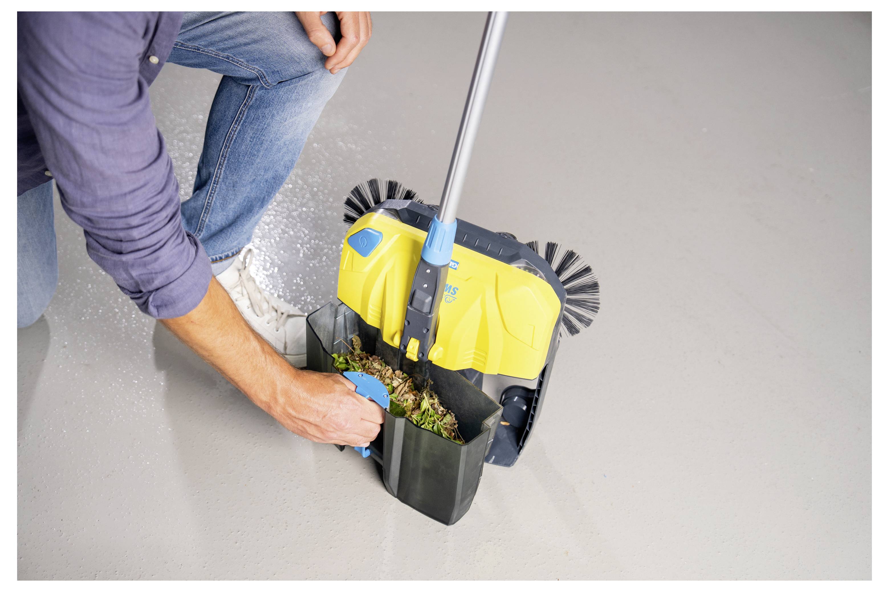 Une personne accroupie sur le sol utilise un petit balai manuel pour ramasser les débris, démontrant la fonctionnalité de l'outil à des fins de nettoyage.