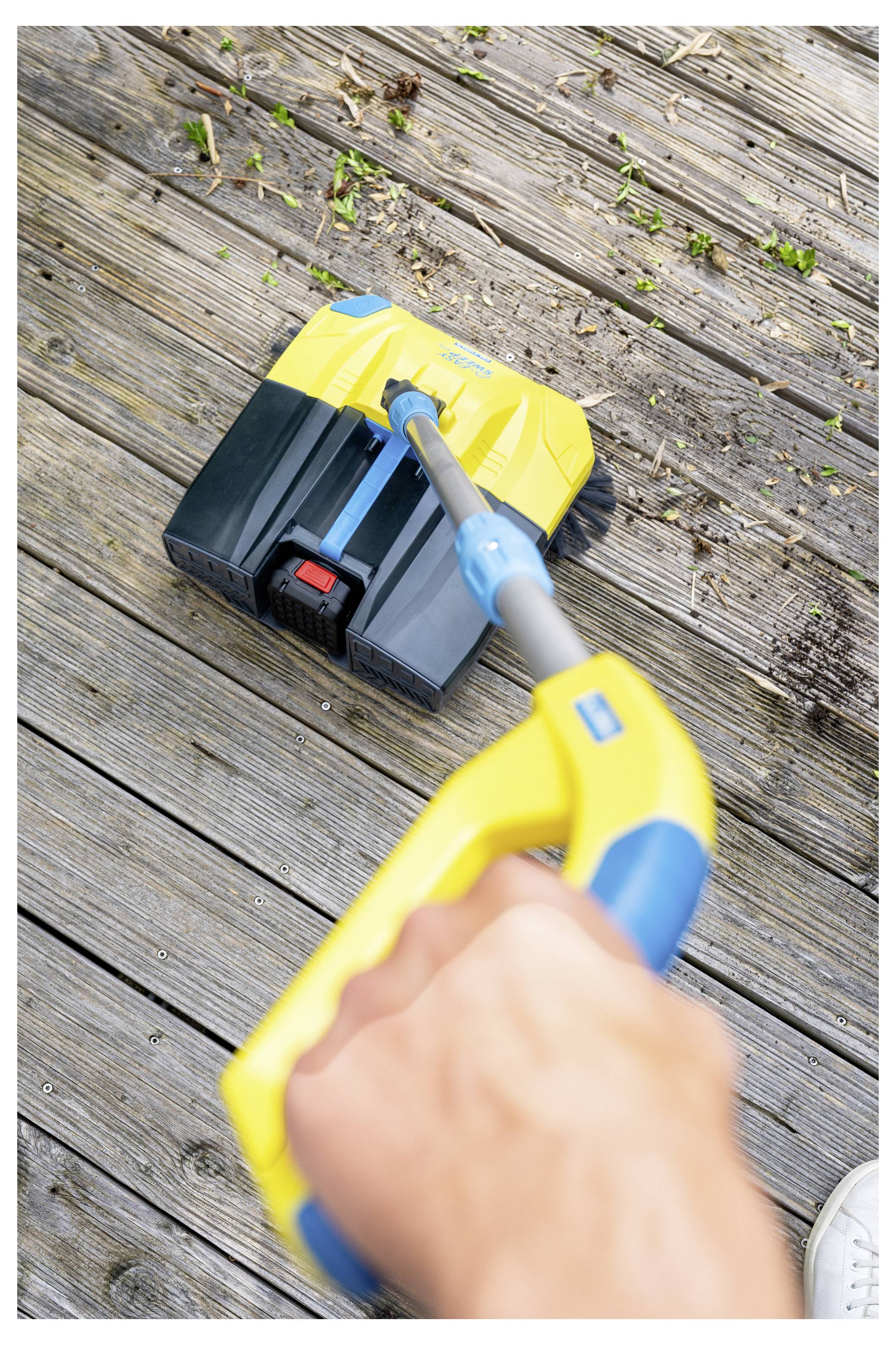 Une main tenant un appareil de nettoyage jaune et bleu frotte un pont en bois, avec de petits débris éparpillés autour.