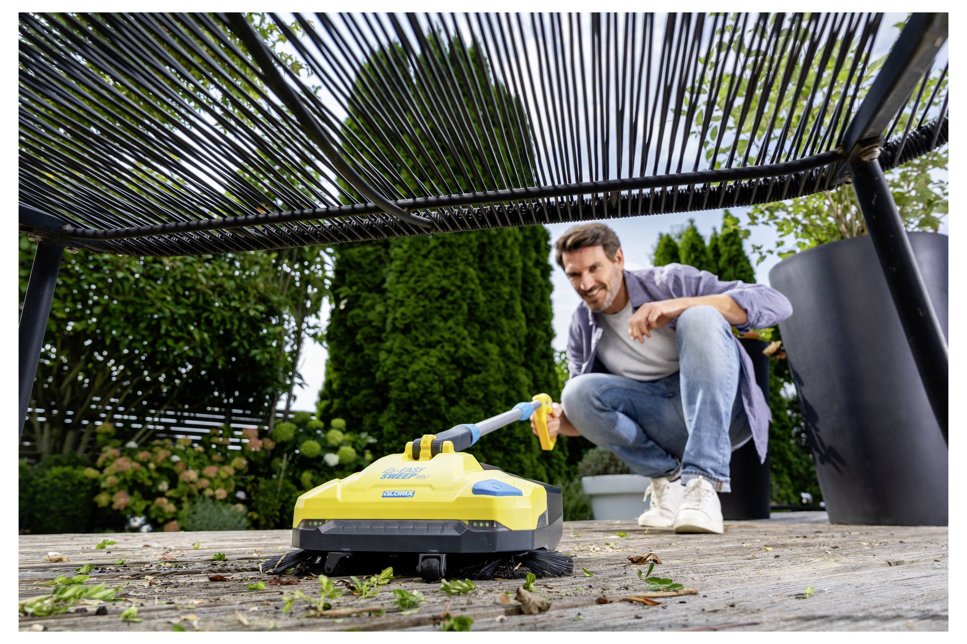 Une personne utilise un aspirateur extérieur jaune pour nettoyer les feuilles d'une terrasse sous une pergola. De la verdure luxuriante et de grands bacs à fleurs se trouvent en arrière-plan.