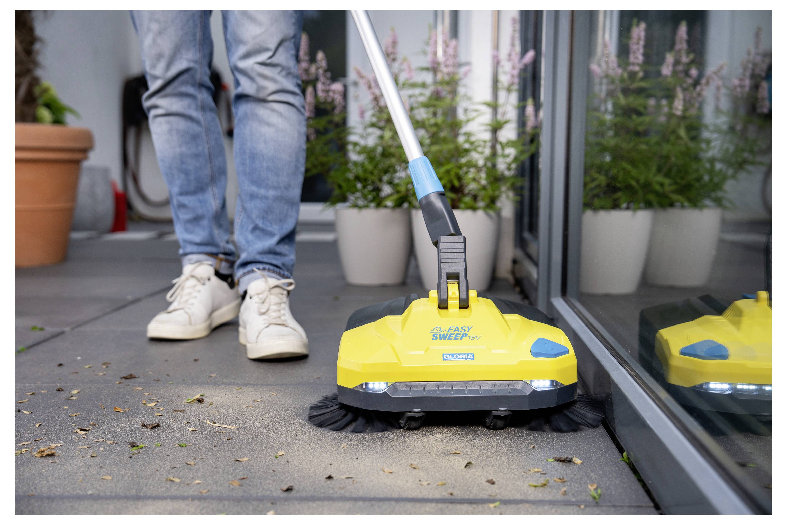 Une personne en jean et baskets utilise un balai-pousseur manuel jaune sur une terrasse, dégageant les débris près des plantes.