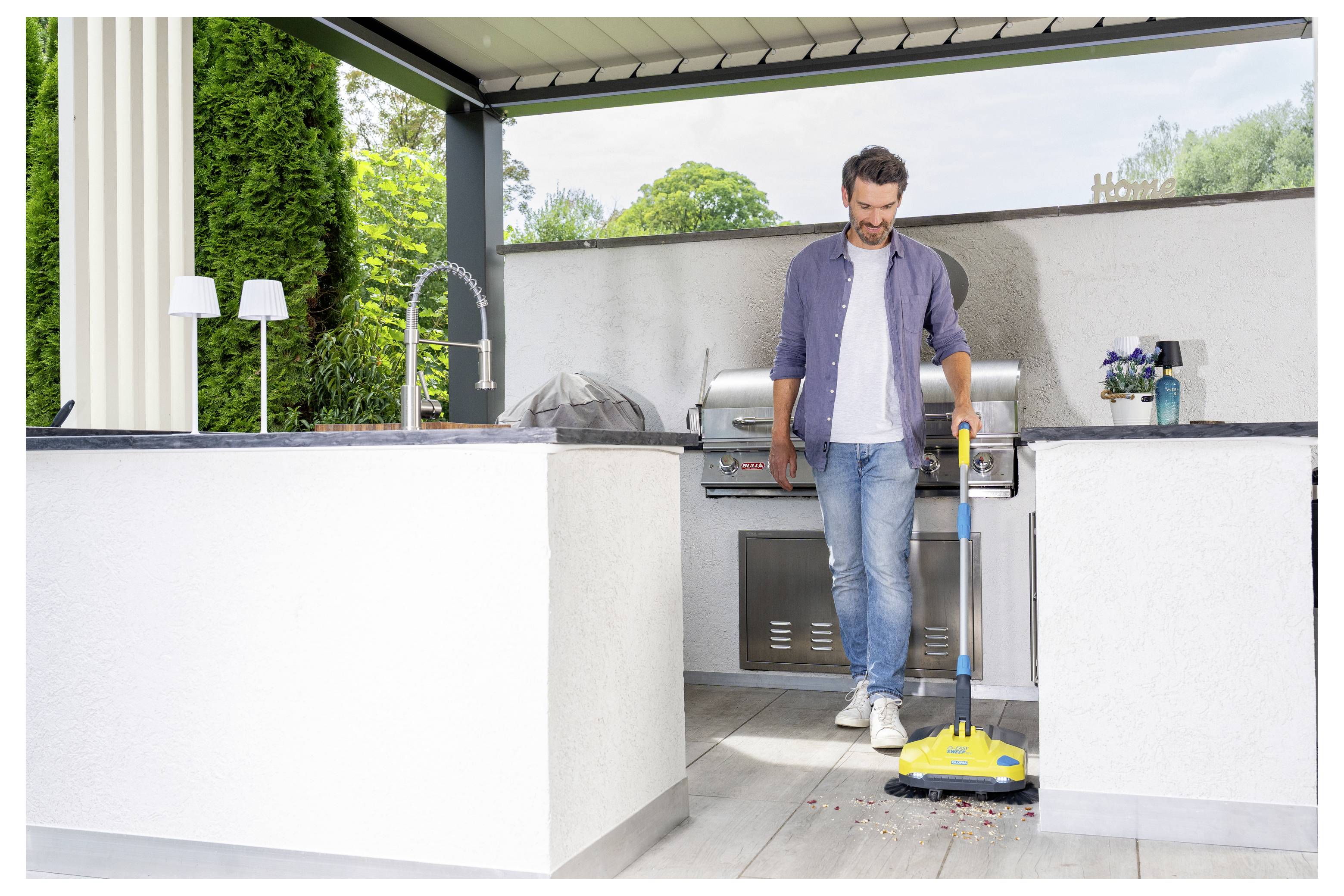Un homme en tenue décontractée utilise un balai jaune pour nettoyer la terrasse d'une cuisine extérieure, avec un barbecue au fond.