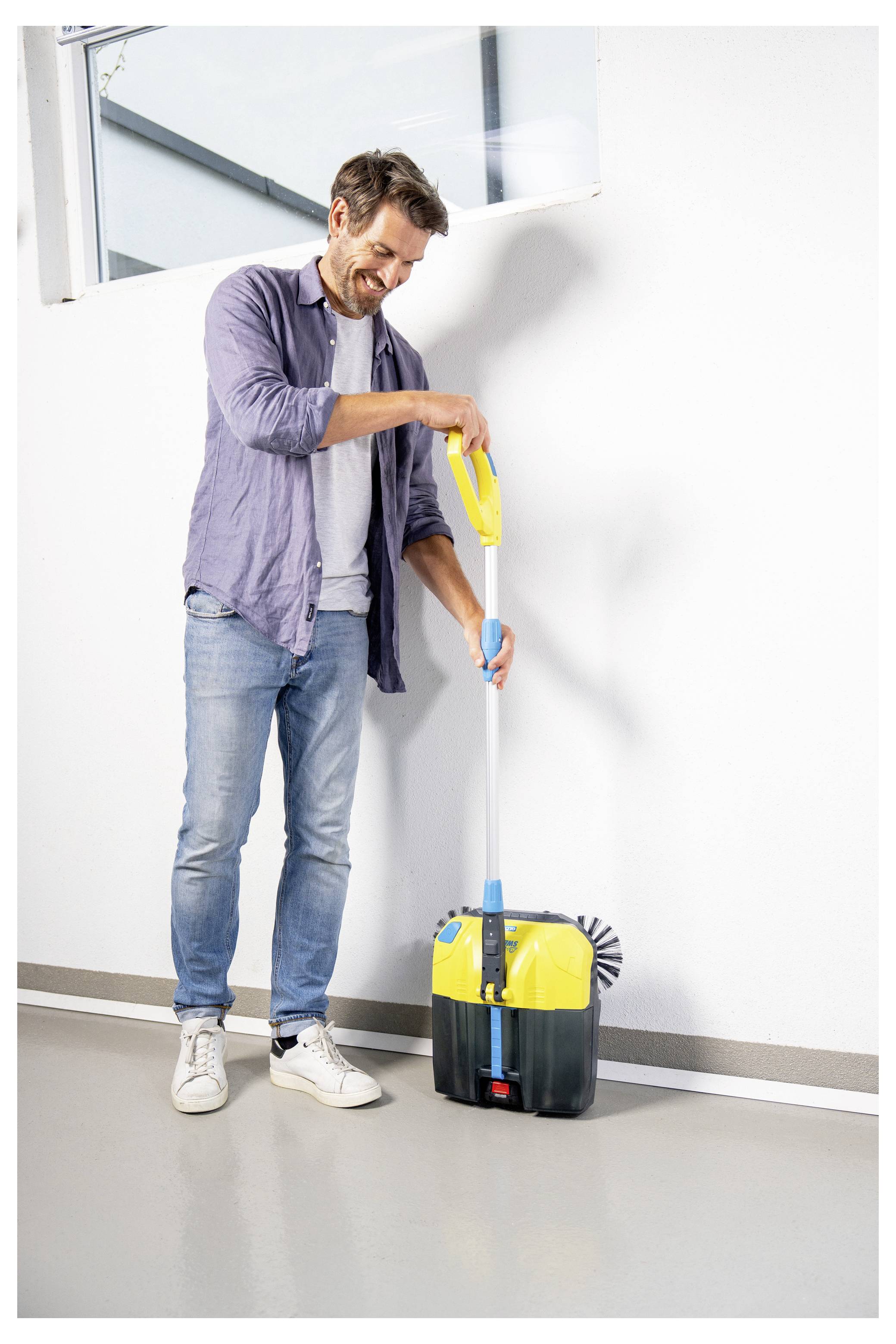 Un homme démontrant un appareil de nettoyage avec une pièce jointe de balai-éponge contre un mur blanc. L'appareil est jaune et noir.