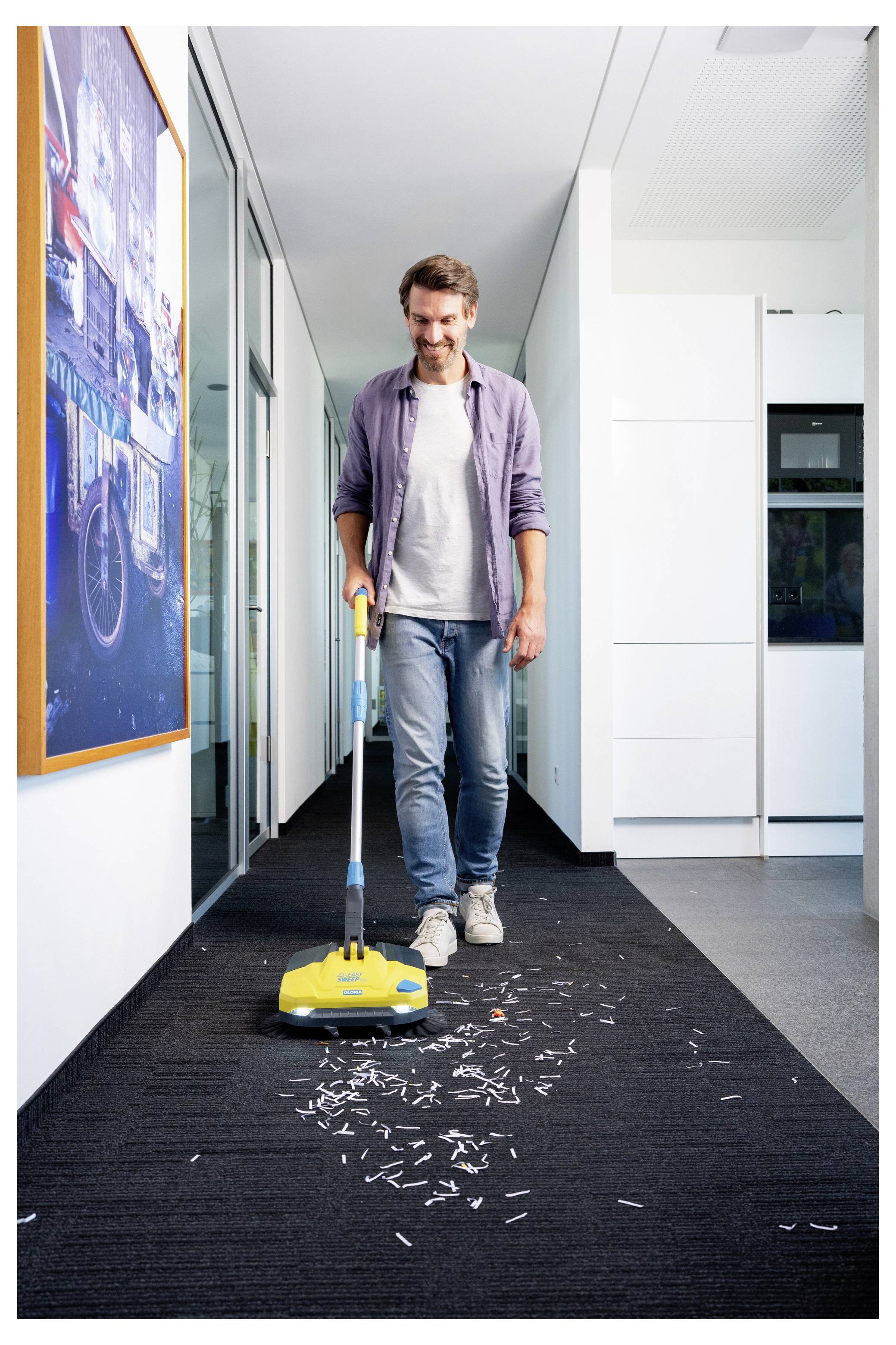 Une personne sourit en passant l'aspirateur sur des confettis éparpillés dans un couloir de bureau moquetté, avec de l'art moderne sur le mur et un environnement propre et lumineux.