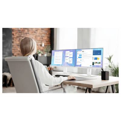 Une femme est assise à un bureau avec deux moniteurs affichant des documents et des applications, dans un bureau à domicile moderne bien éclairé.