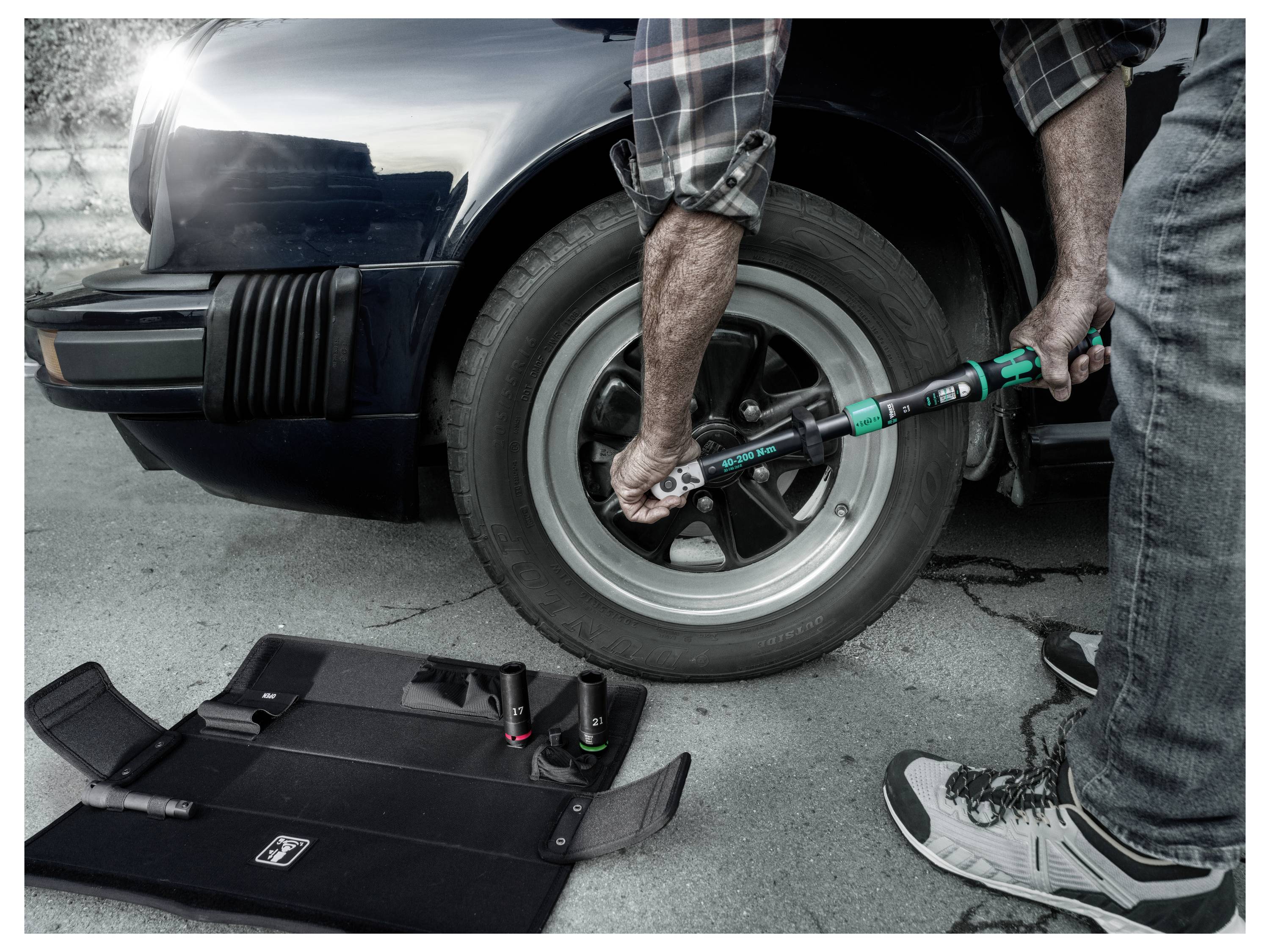 Une personne utilise une clé dynamométrique pour serrer un écrou de roue sur une roue de voiture, avec une mallette à outils ouverte sur le sol.