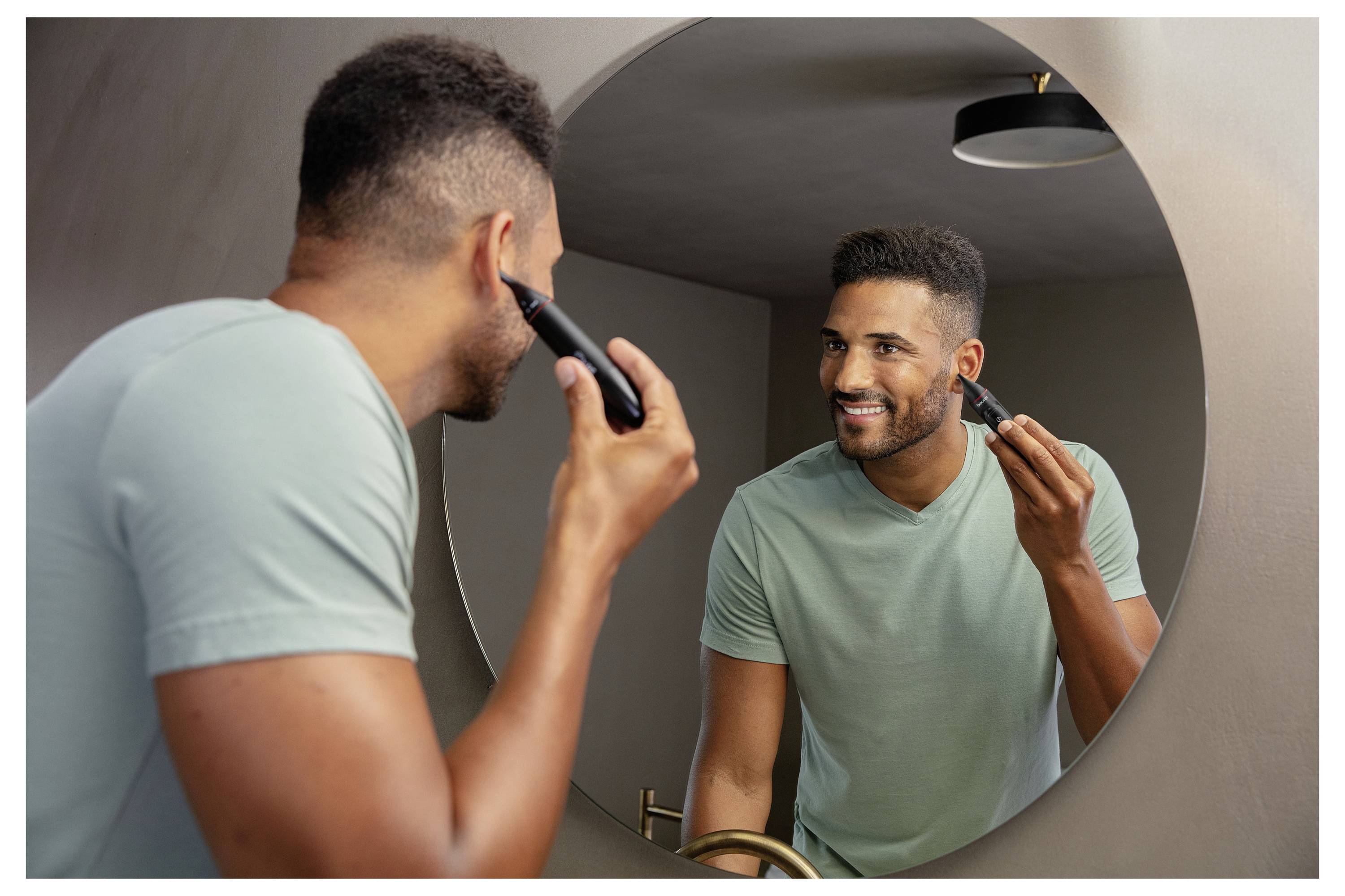 Un homme en chemise verte sourit en utilisant un rasoir électrique devant un miroir de salle de bain circulaire.