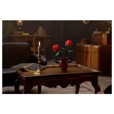 Une pièce faiblement éclairée avec une table en bois, présentant une bougie allumée, un livre ouvert et un vase de roses rouges. Des bagages vintage en arrière-plan.