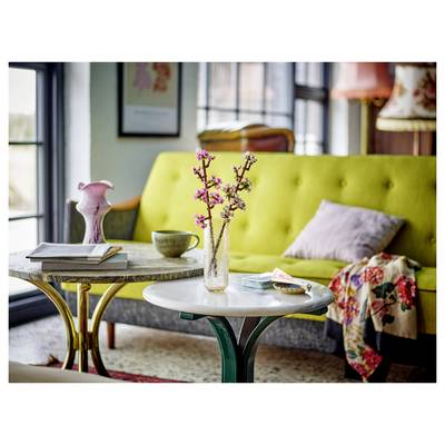 Un vase avec des fleurs de cerisier sur une petite table, disposé dans un salon avec un canapé jaune, un coussin rose et des objets décoratifs.