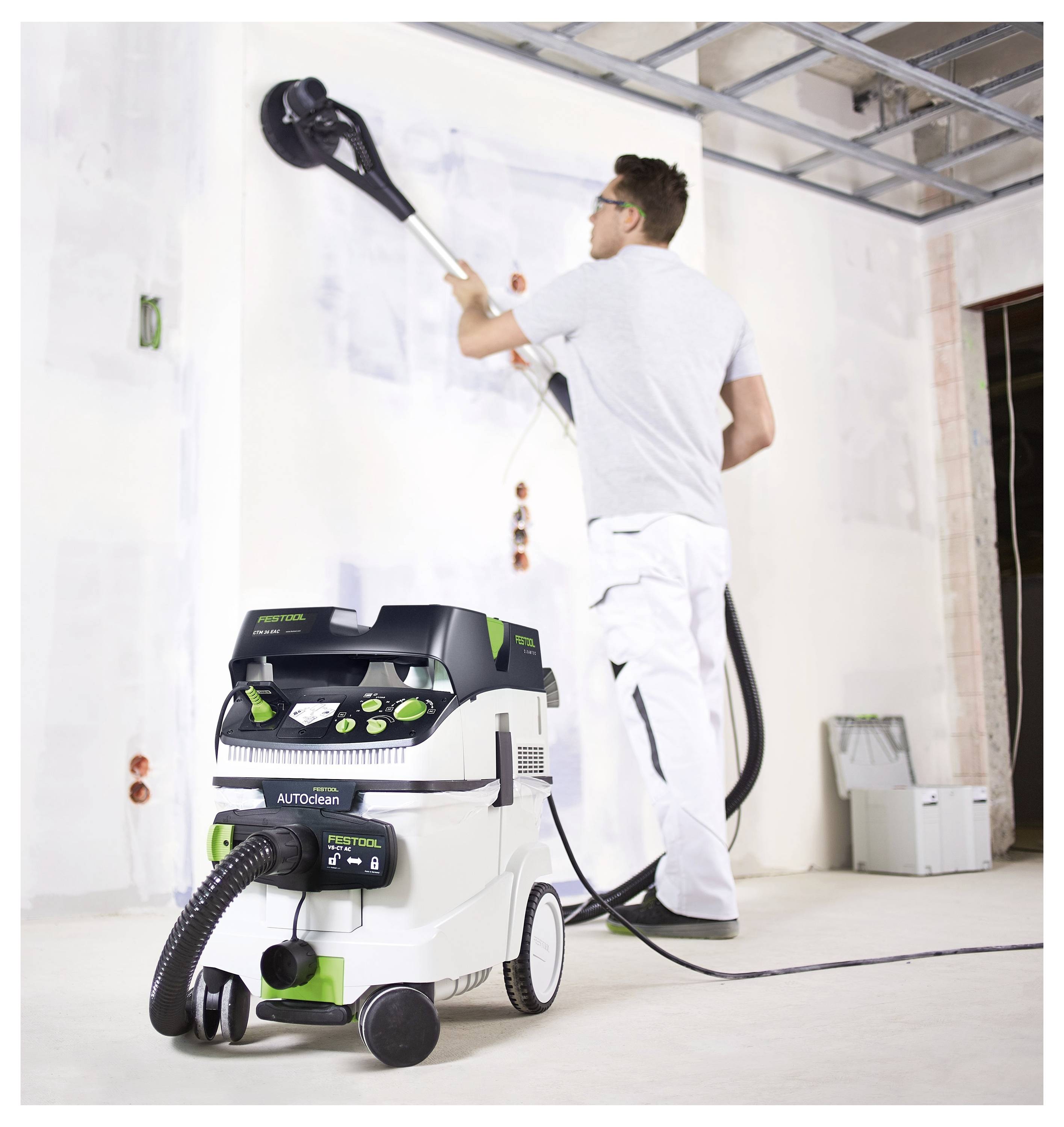 Une personne vêtue de tenue de travail utilise un outil de ponçage mural connecté à un système d'aspiration dans une pièce en construction, en se concentrant sur le lissage des murs.