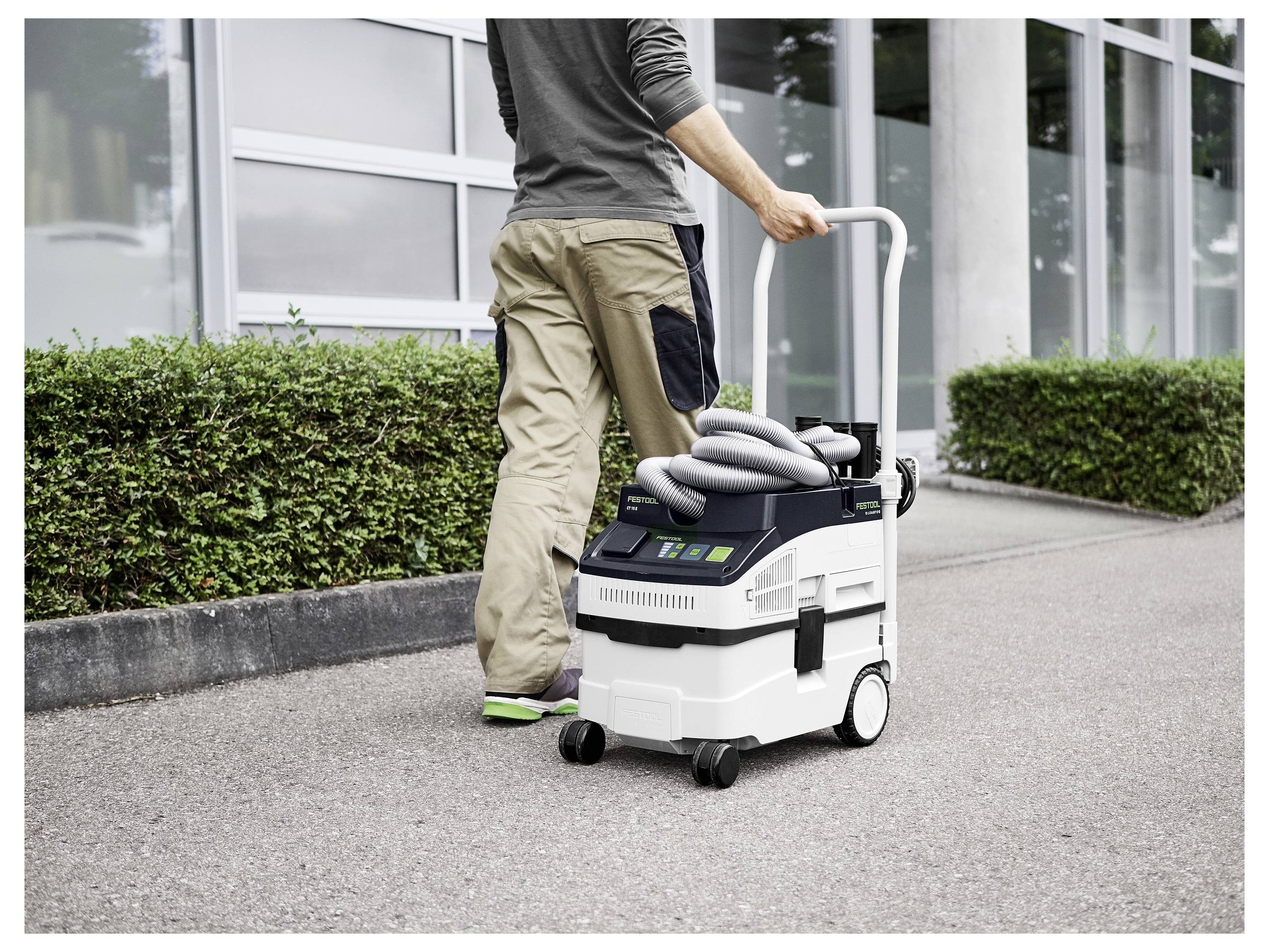 Une personne pousse un aspirateur portable avec tuyau enroulé sur un trottoir près d'un bâtiment, mettant en avant l'utilisation en extérieur et la mobilité.