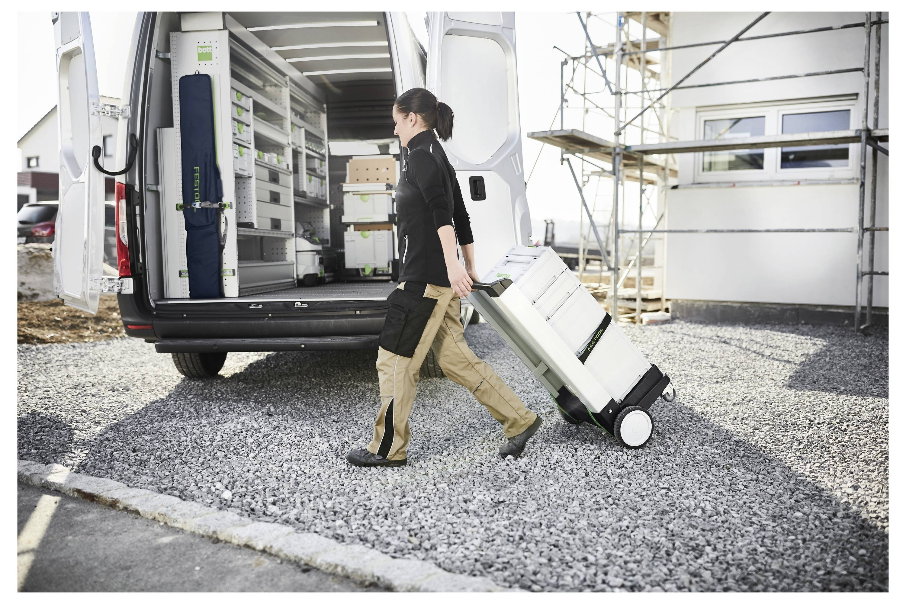 Une personne tire une boîte à outils à roulettes hors d'une camionnette garée sur un chantier de construction résidentiel, ce qui suggère l'organisation et la préparation dans l'environnement de travail.