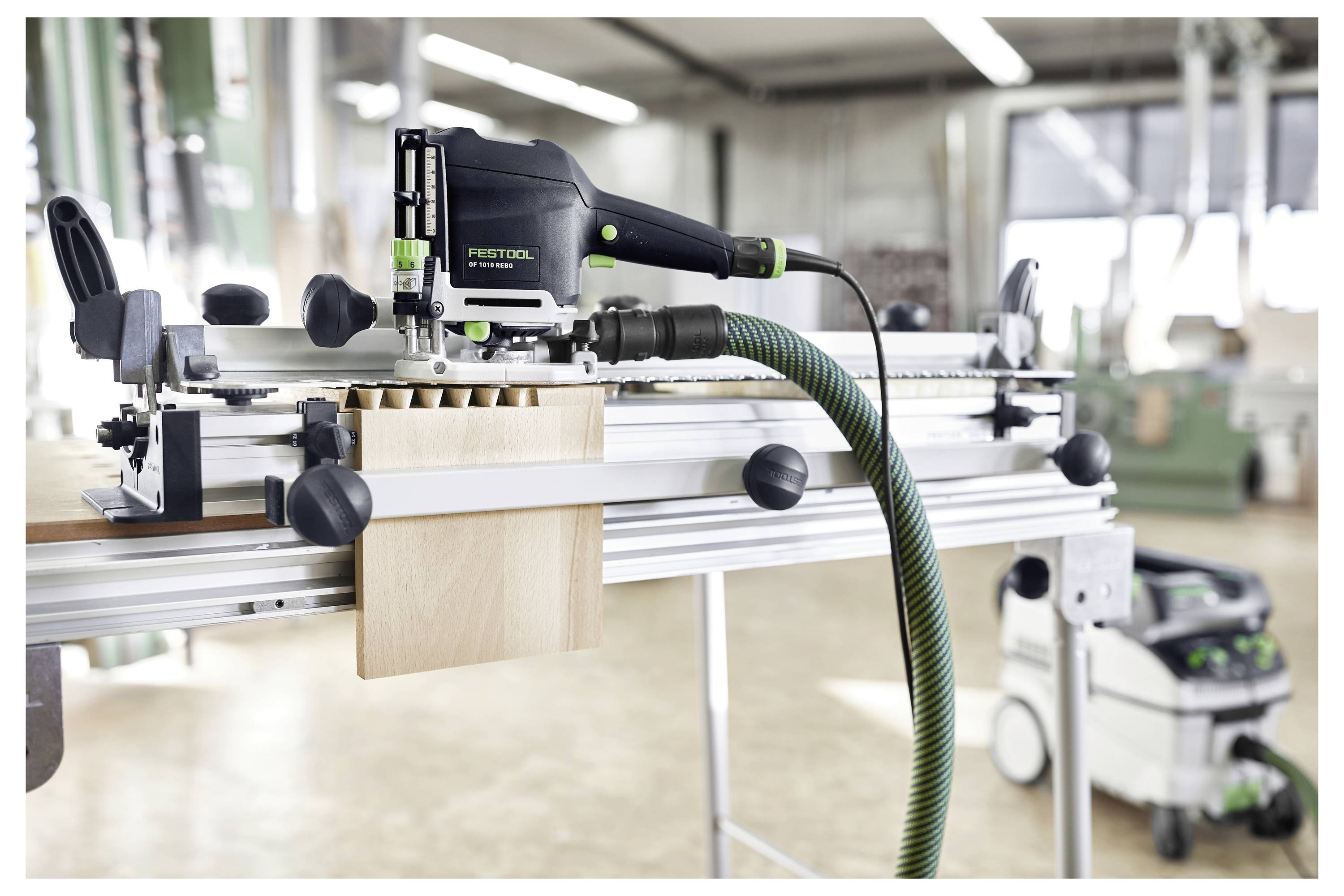 Scène d'atelier affichant un outil Festool monté sur un établi, apparemment utilisé pour le travail du bois, avec des accessoires connectés et un aspirateur d'atelier en arrière-plan.