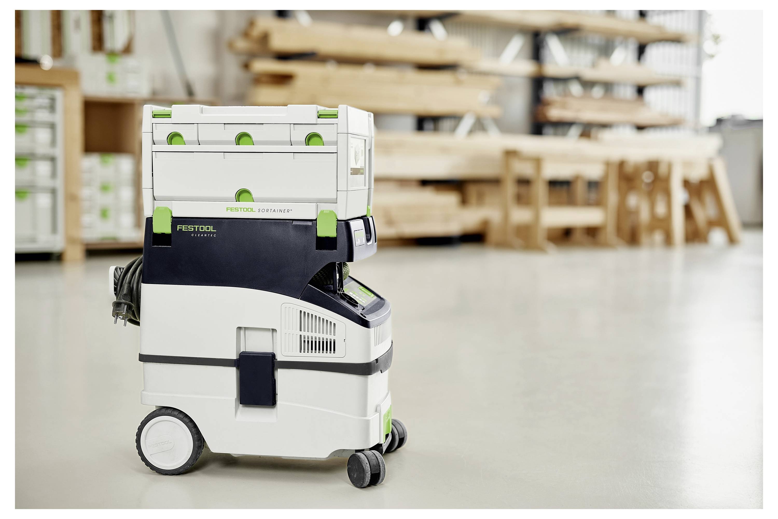 Un extracteur de poussière Festool se trouve au sol d'un atelier de menuiserie, avec des établis en bois et des étagères de rangement flous en arrière-plan.