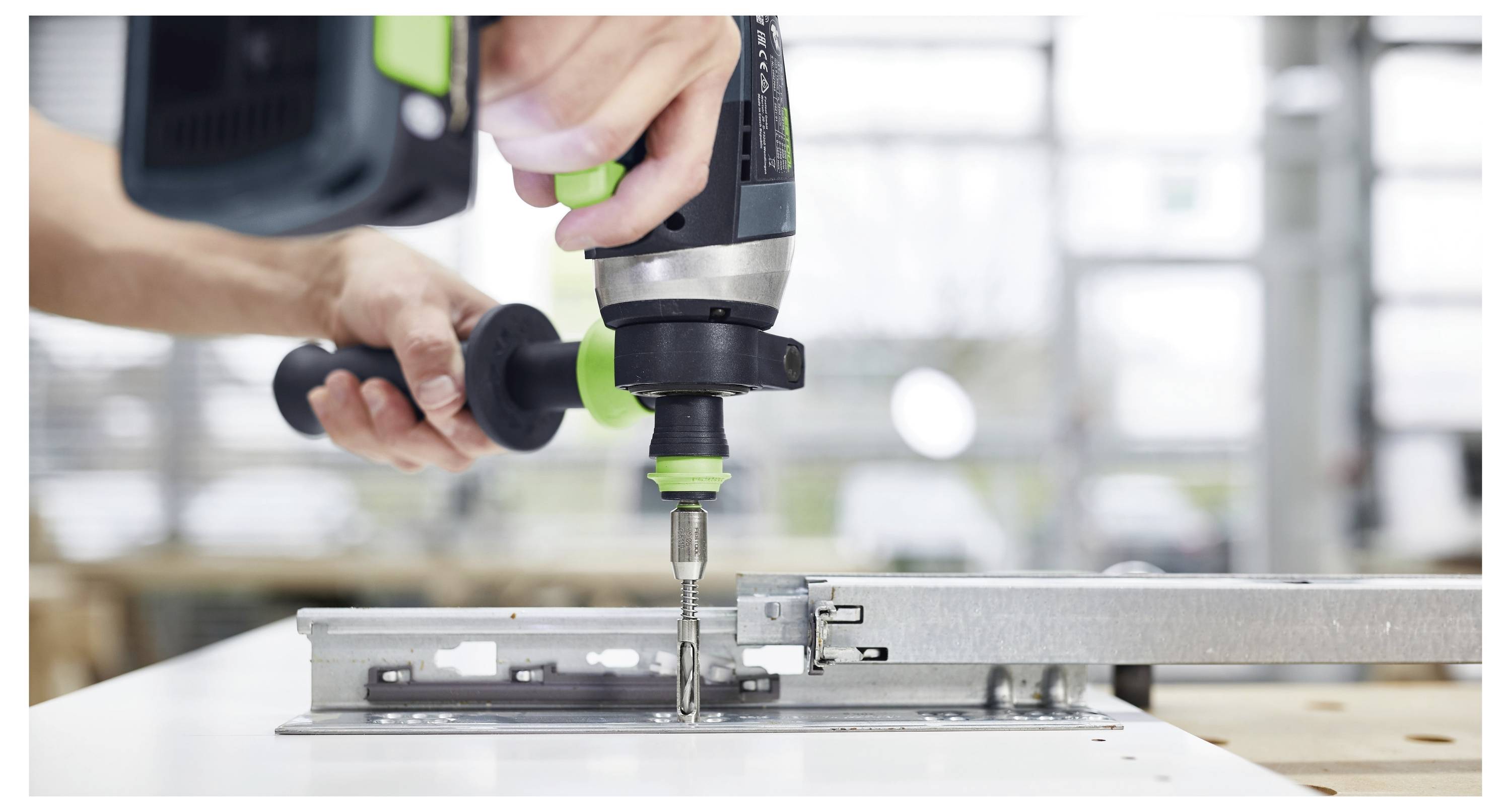 Une personne utilisant une perceuse électrique pour fixer un support métallique à une surface blanche dans un atelier.