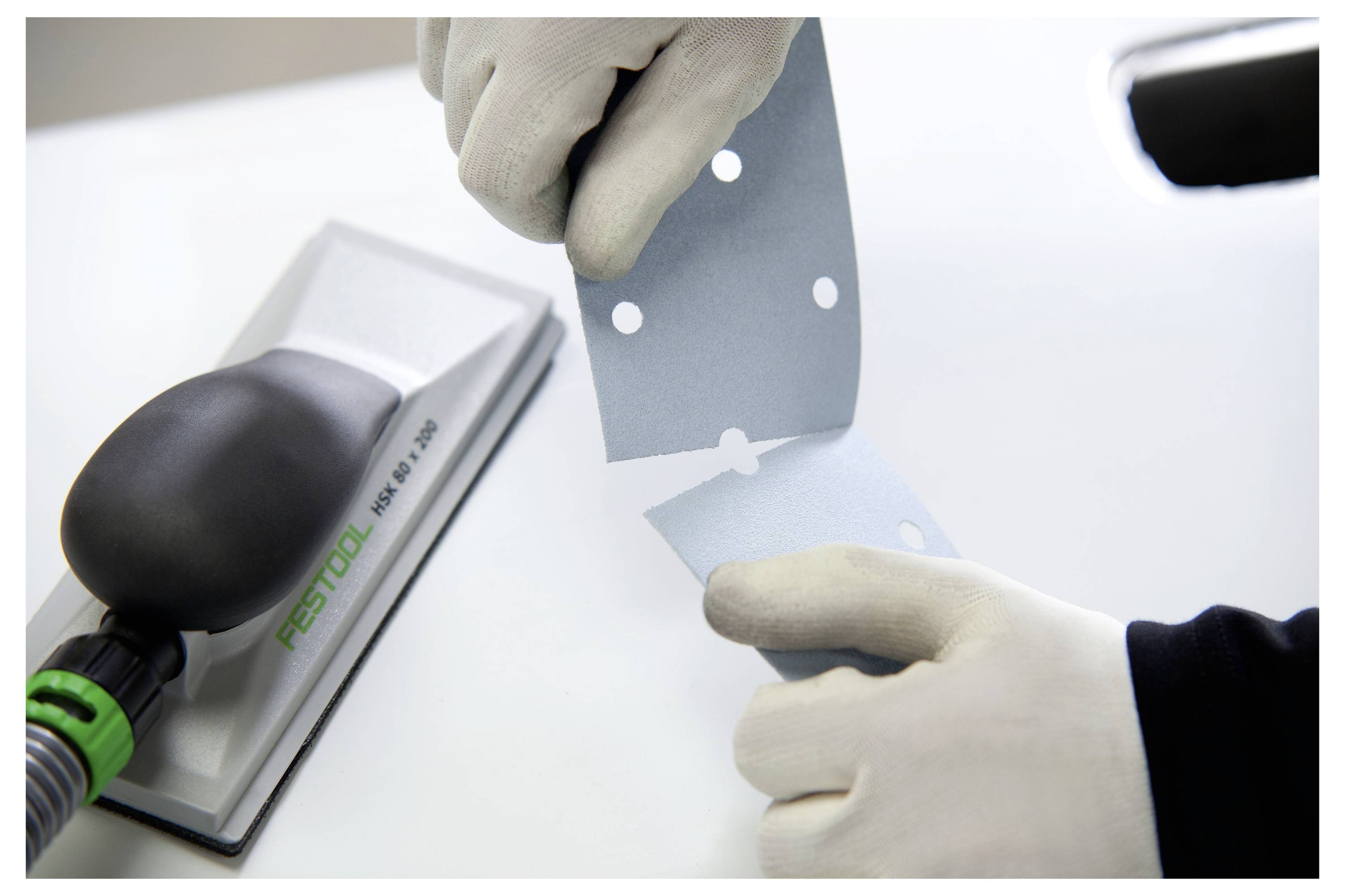 Une personne portant des gants déchire une feuille de papier de verre bleu avec des trous pré-découpés à côté d'un outil de ponçage Festool sur une surface blanche.